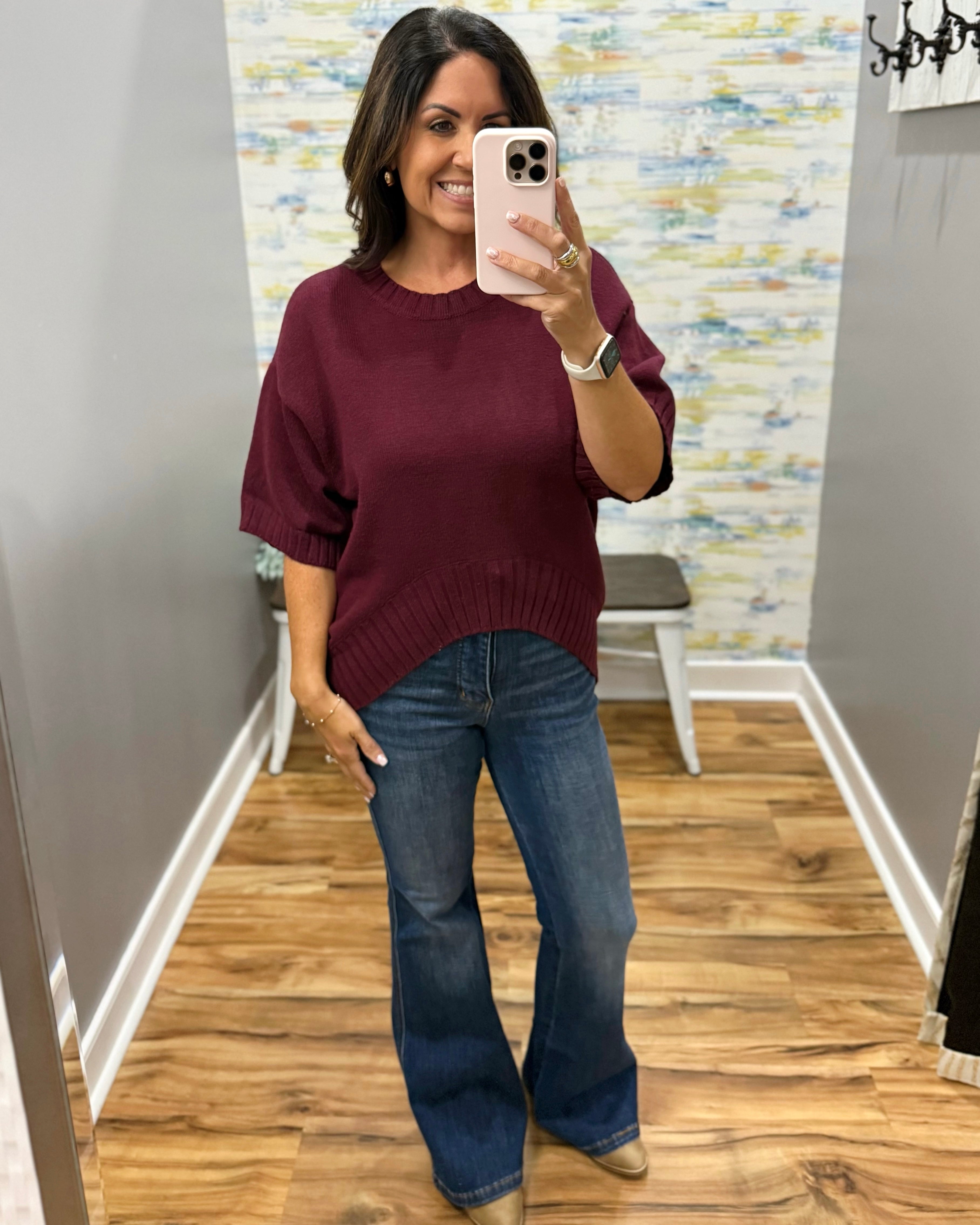 A woman takes a mirror selfie in a fitting room wearing a loose-fitting, short-sleeve crewneck sweater in burgundy and blue flare jeans.