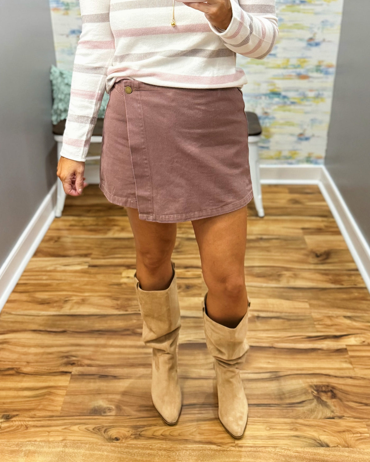 A person wearing a chestnut color wrap-style mini skirt with a single button closure, paired with a pink and white striped long-sleeve top and Corkys City Slicker boots in Sand Faux Suede.