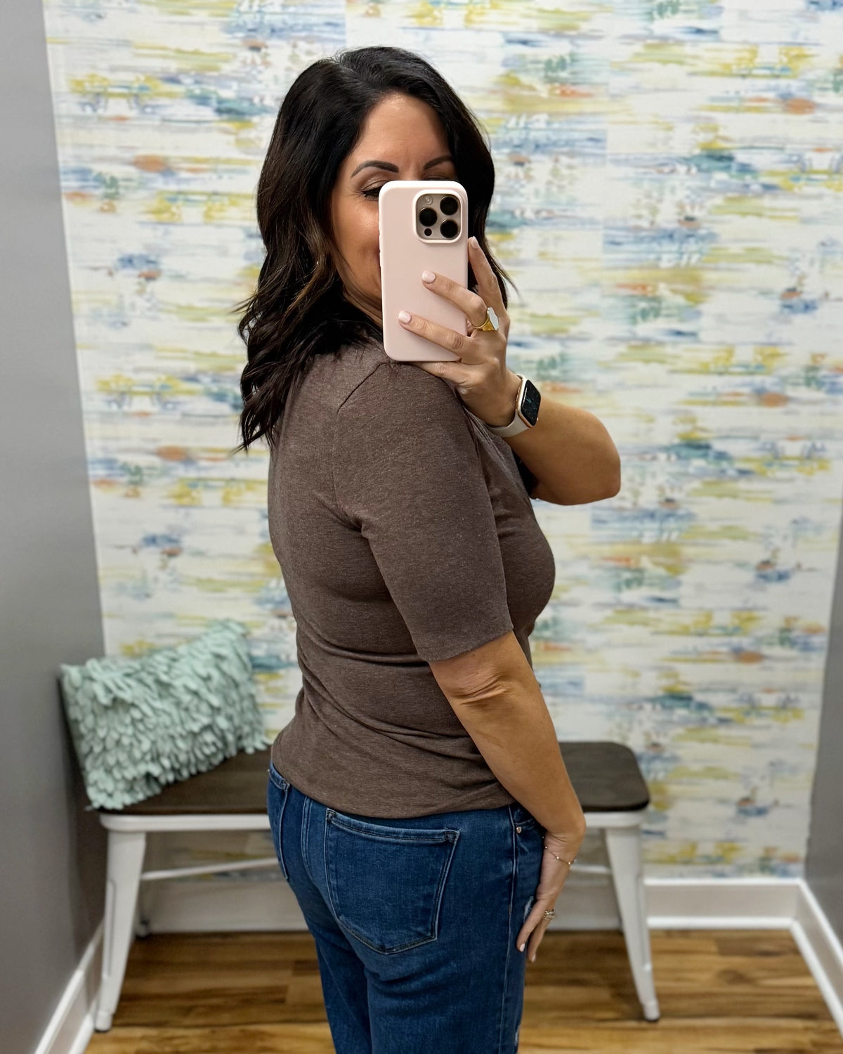A woman takes a mirror selfie in a fitting room wearing a fitted short-sleeve brown top and blue jeans, with patterned wallpaper and a bench in the background.