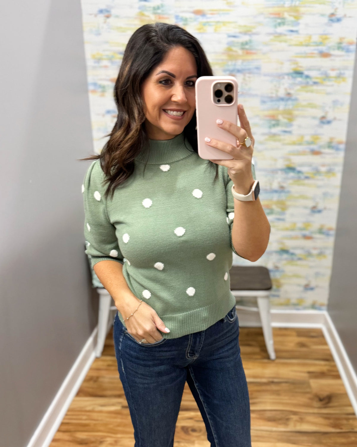 A woman takes a mirror selfie wearing a sage green mock-neck knit sweater with white polka dots and dark blue jeans in a boutique fitting room.