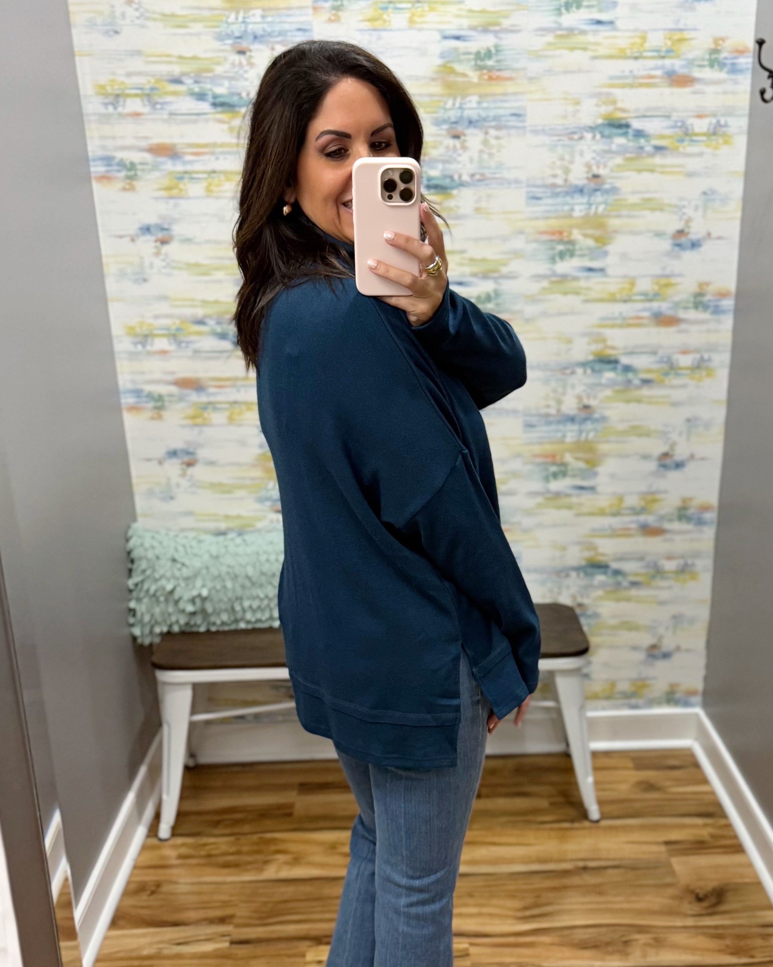 A woman takes a mirror selfie in a fitting room wearing an oversized dark teal blue knit top and light blue jeans.