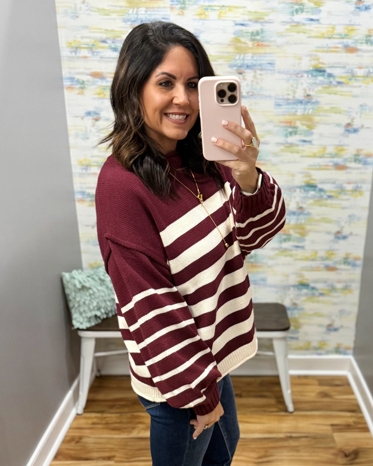 A woman takes a mirror selfie wearing a maroon and cream striped knit sweater, dark blue jeans, and a gold necklace. The background shows a gray wall, a white bench with a green pillow, and a colorful abstract patterned wallpaper.