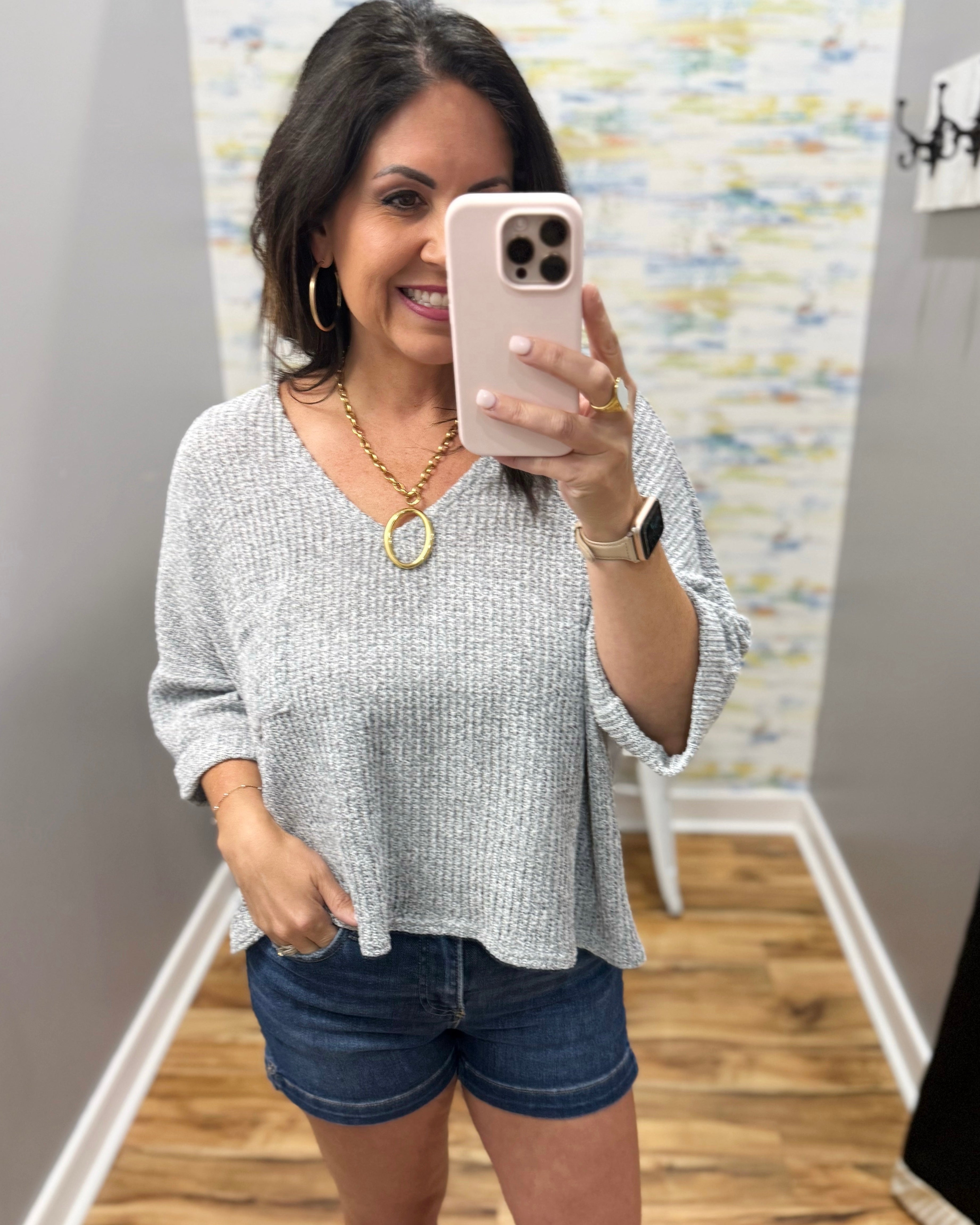 Woman taking a mirror selfie wearing a gray sweater and denim shorts in a room with a colorful wall.