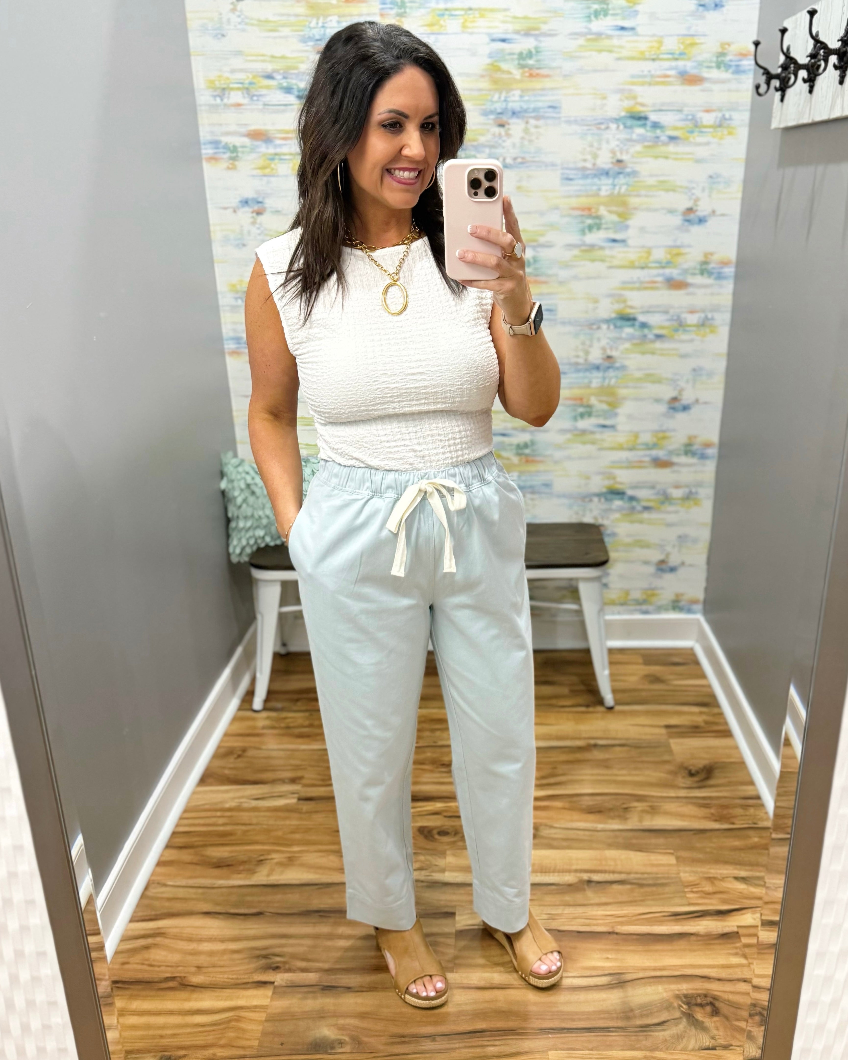 Woman wearing a white top and light gray pants in a room with a patterned wall.