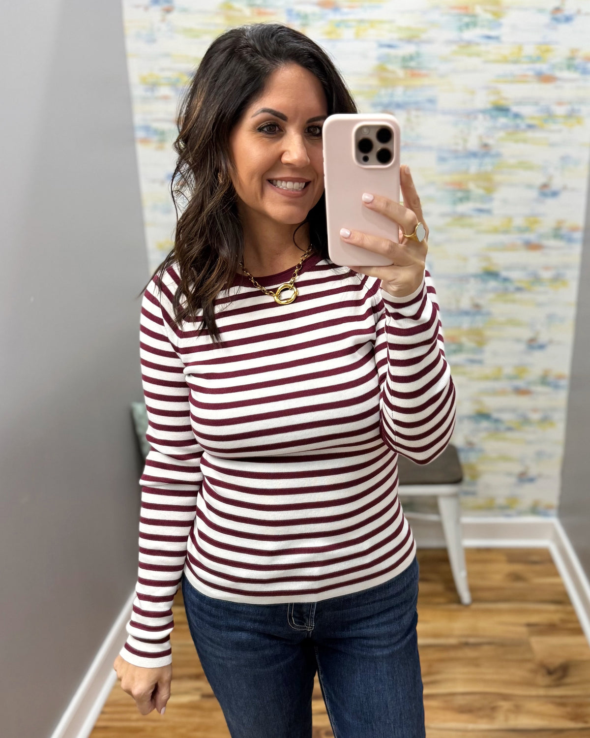A woman taking a mirror selfie wearing a long-sleeve crewneck top with horizontal burgundy and white stripes, paired with dark blue jeans.