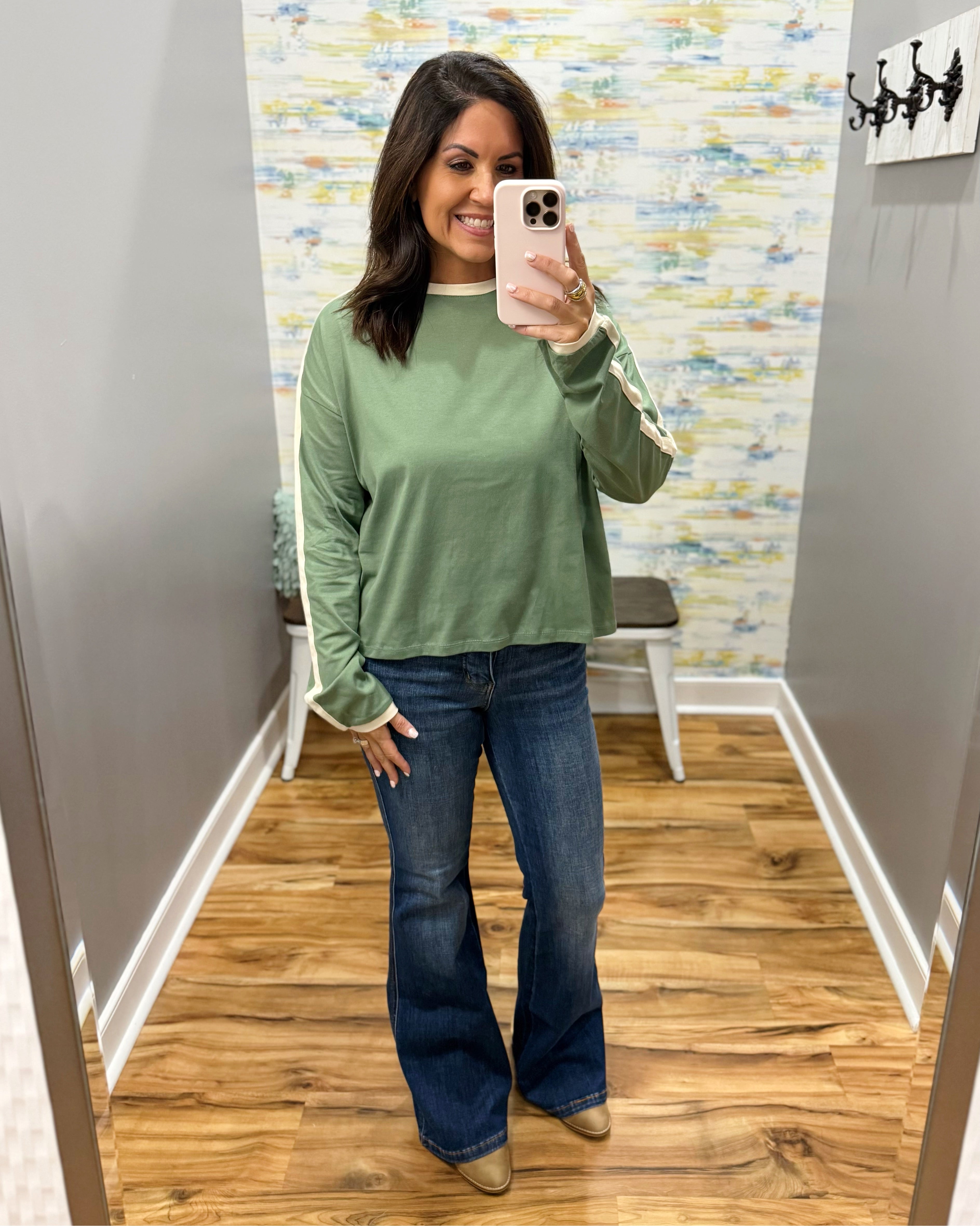 A woman takes a mirror selfie in a fitting room wearing a sage green long-sleeve top with white ribbed trim, blue flare jeans, and tan boots.