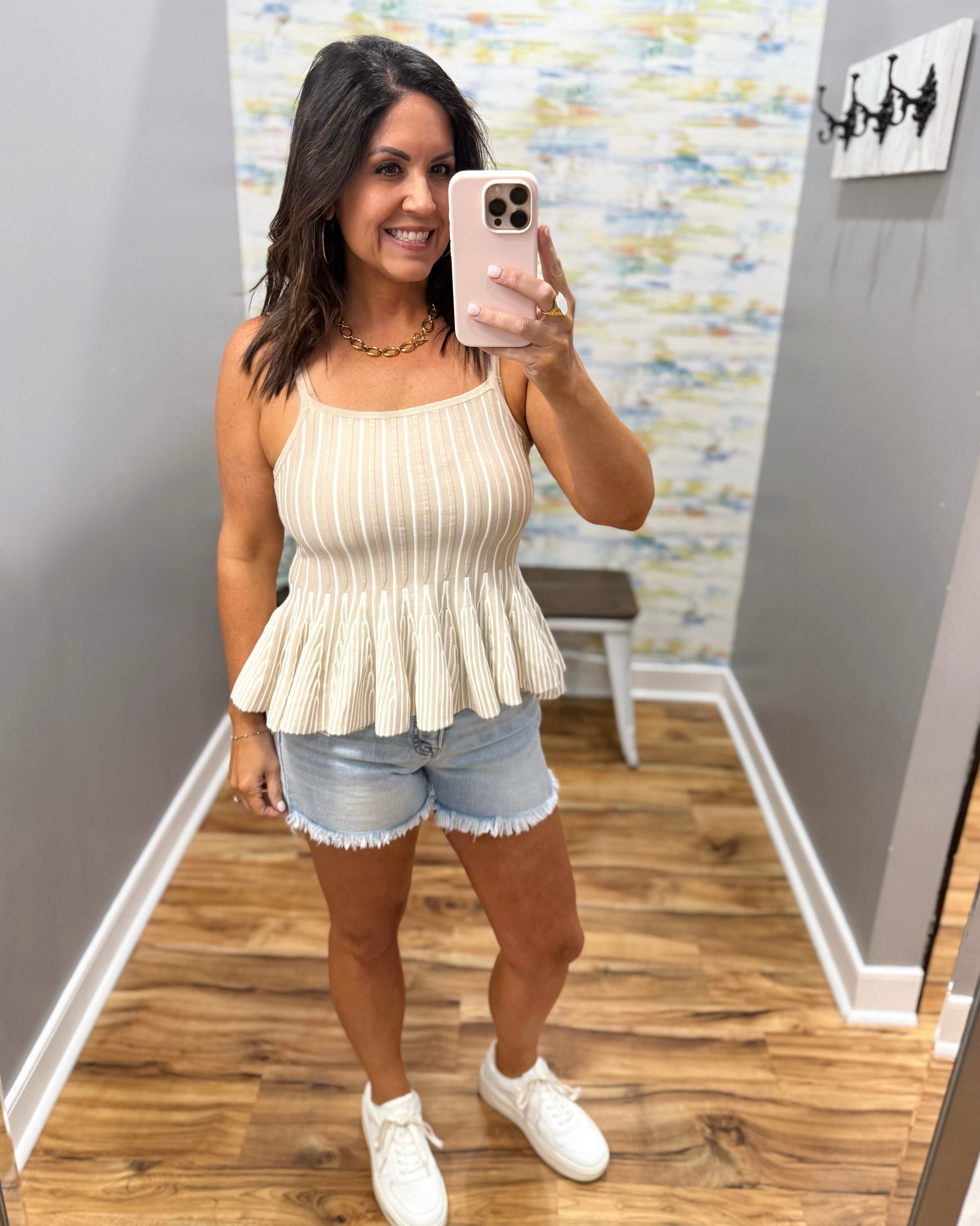 Woman taking a mirror selfie wearing a beige peplum top and denim shorts in a room with a colorful wall.