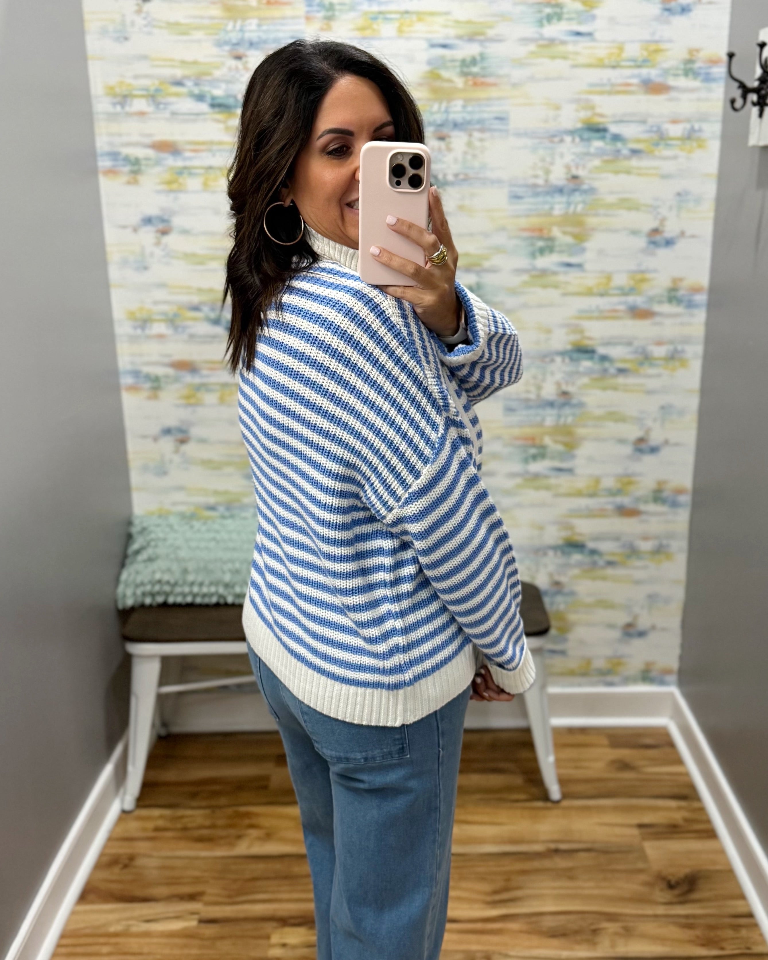 A woman takes a mirror selfie in a fitting room wearing a blue and white striped knit cardigan sweater and blue jeans.