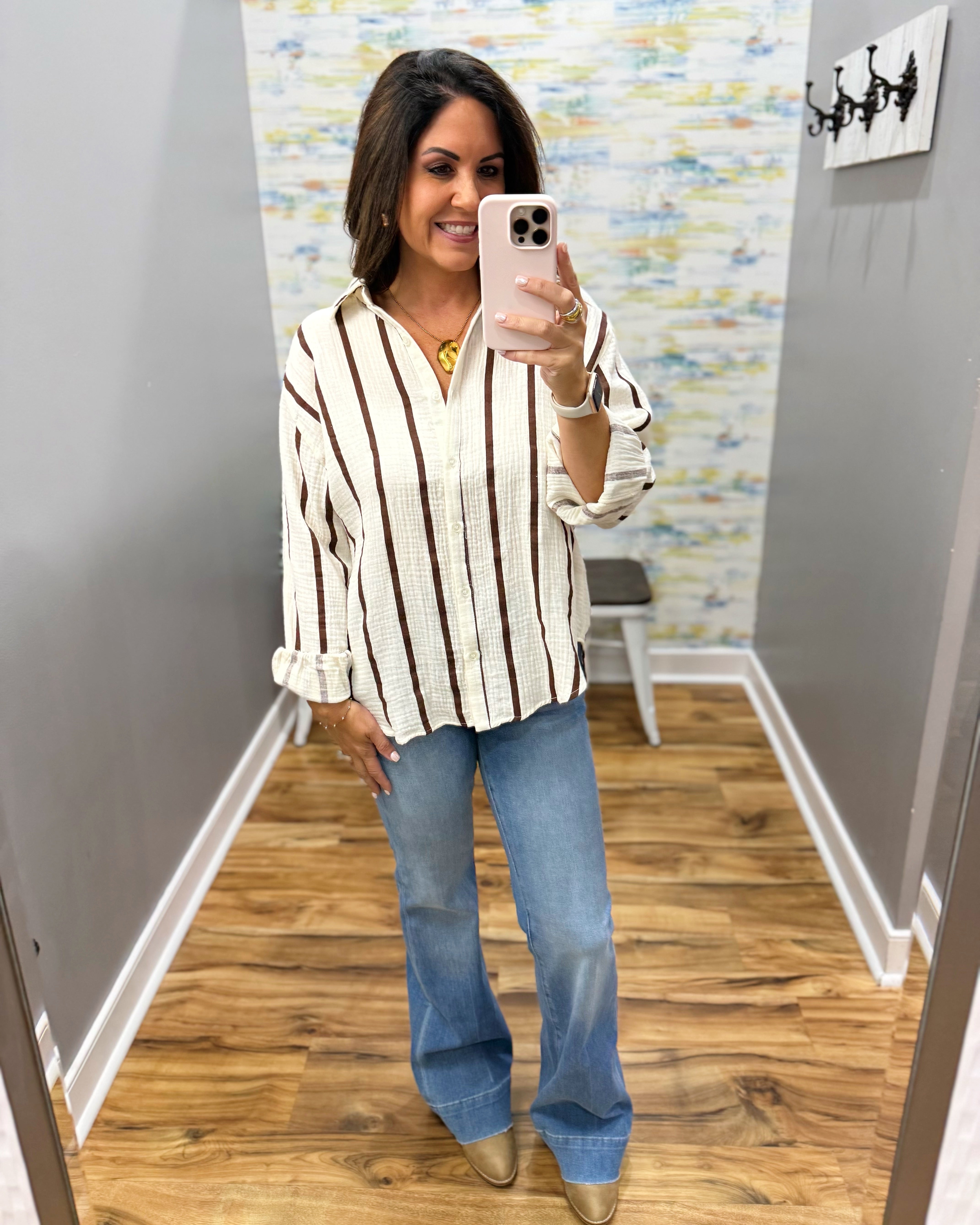 A woman takes a mirror selfie in a fitting room wearing a cream and brown vertically striped button-down shirt, blue flare jeans, and metallic gold pointed-toe flats.