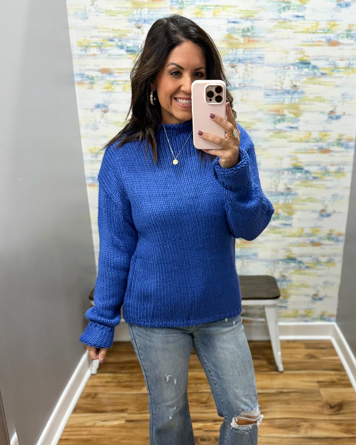A woman takes a mirror selfie wearing a bright royal blue textured knit sweater and light-wash distressed jeans in a fitting room with a patterned wallpaper background.