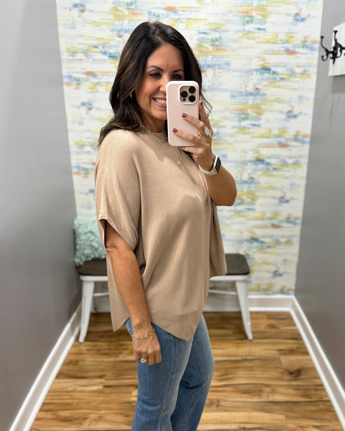 A woman takes a mirror selfie wearing a beige short-sleeve knit top and blue jeans in a room with a patterned wallpaper background.