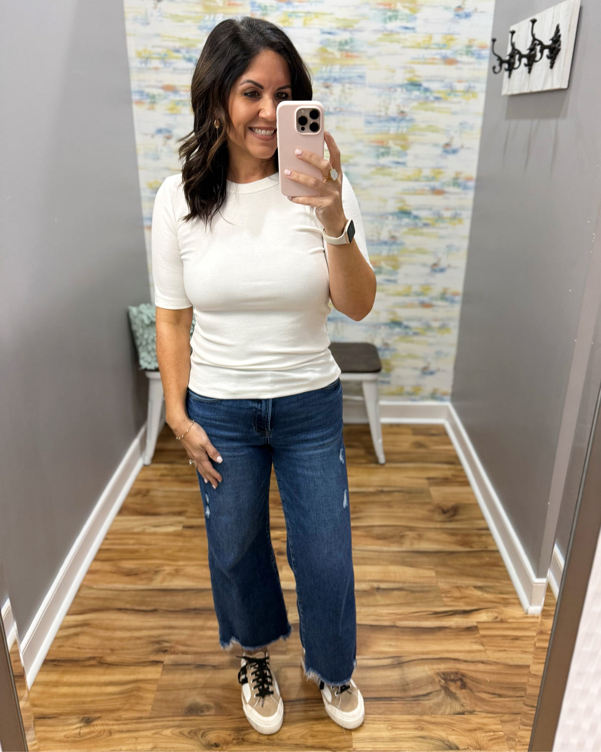 A woman takes a mirror selfie in a fitting room wearing a fitted white short-sleeve top, wide-leg blue jeans, and beige sneakers.