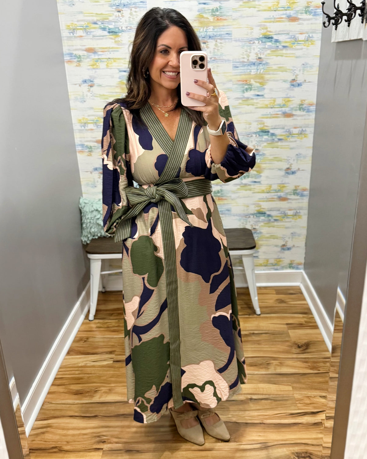 A woman takes a mirror selfie in a fitting room wearing a long-sleeve wrap maxi dress with a green, beige, and navy camouflage pattern. The dress features a tie waist belt and is paired with beige pointed-toe flats.