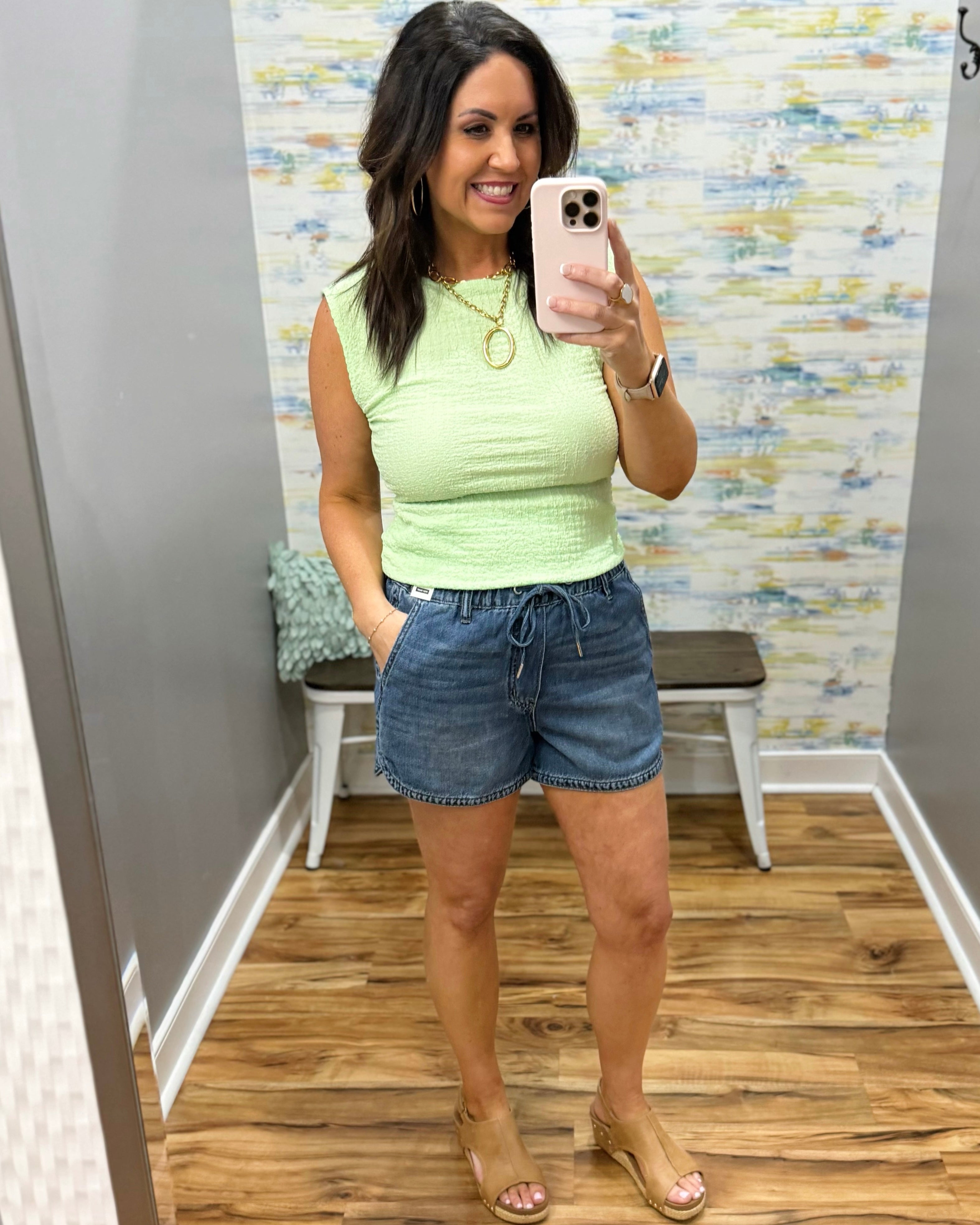 Woman wearing a light green sleeveless top and denim shorts in a room with a colorful wall.