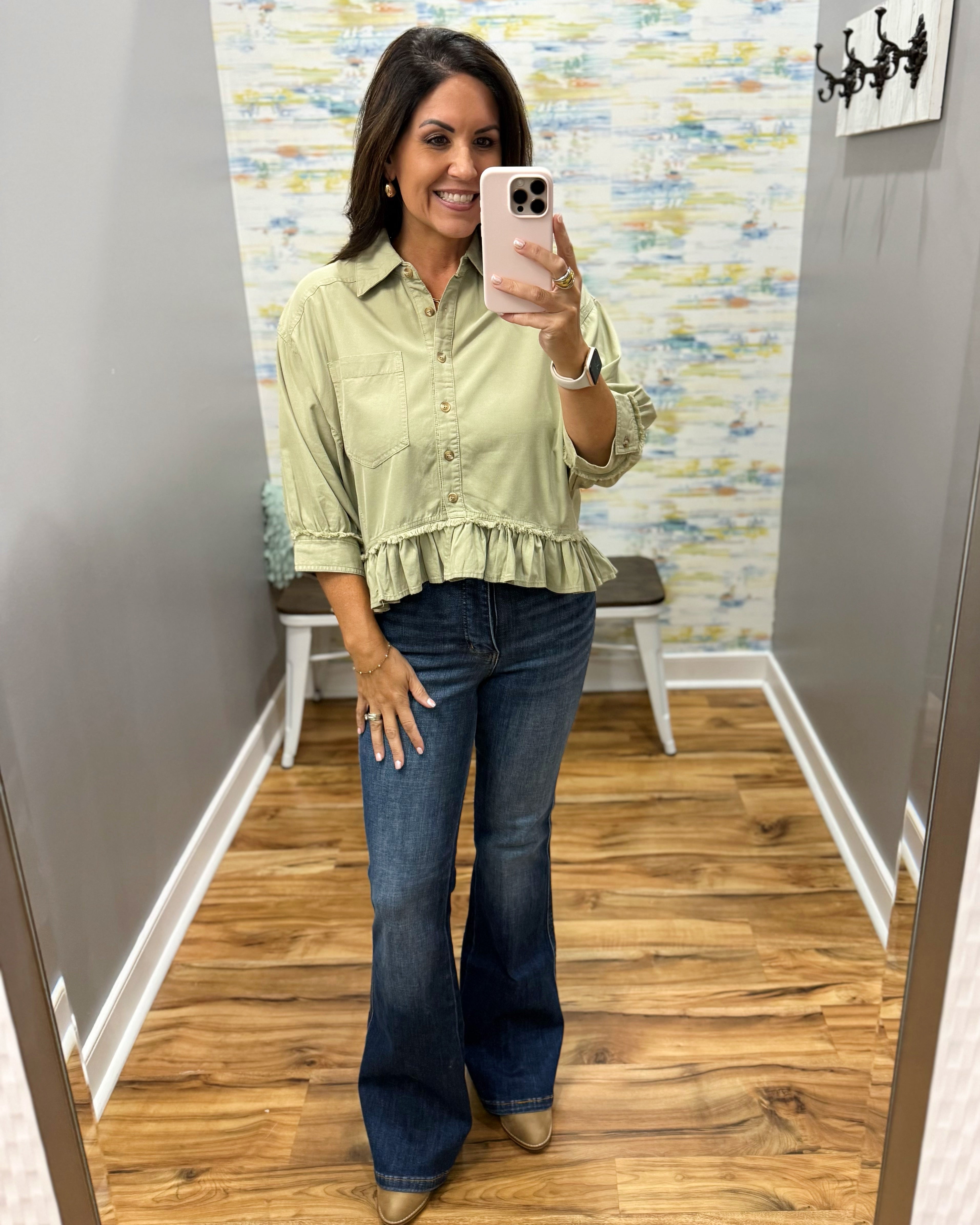 A woman takes a mirror selfie wearing a sage green ruffled button-up blouse and dark blue flare jeans in a fitting room with wood floors and a patterned accent wall.