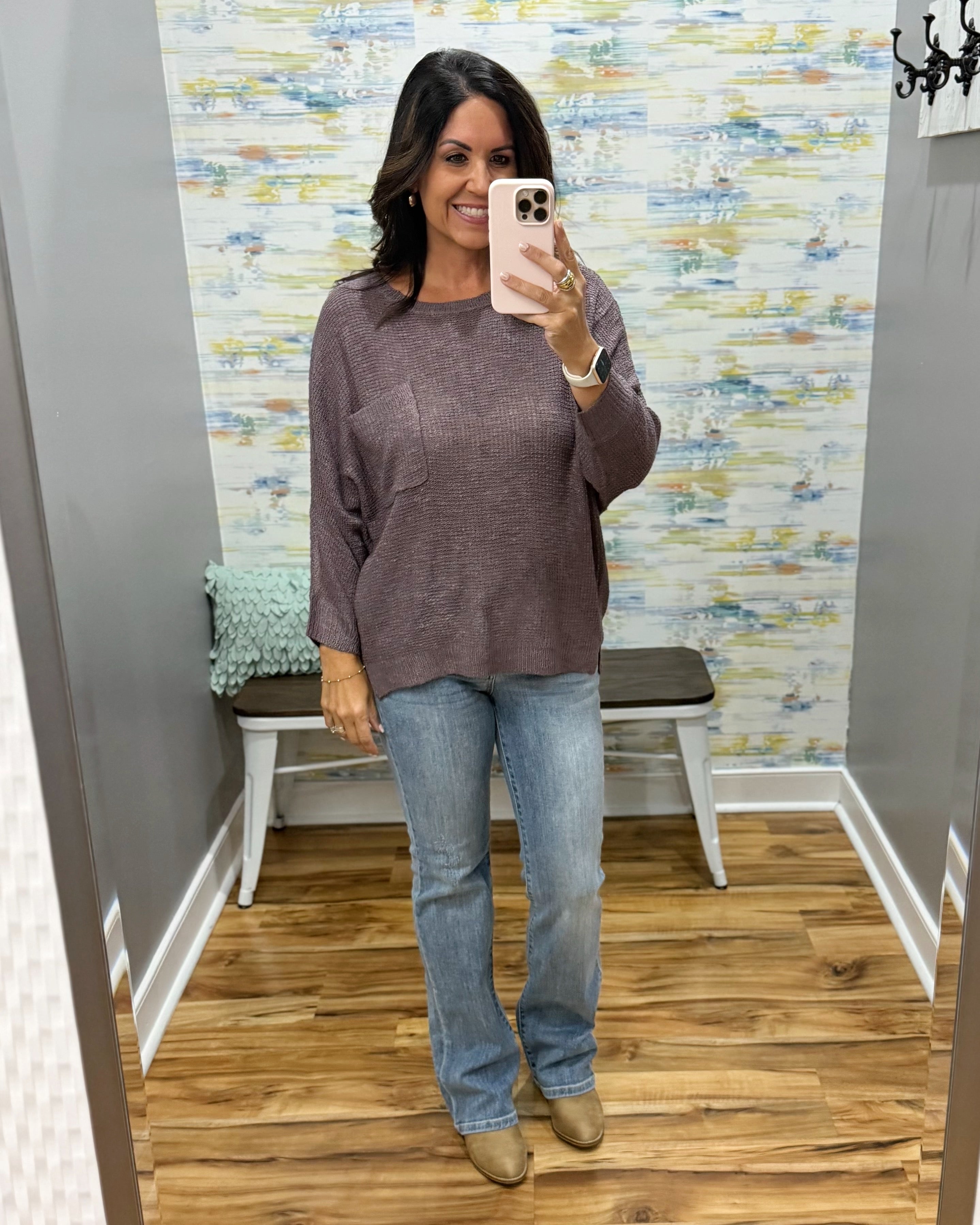 A woman takes a mirror selfie in a fitting room wearing a heathered mauve-brown long-sleeve knit top, light-wash blue jeans, and tan boots.