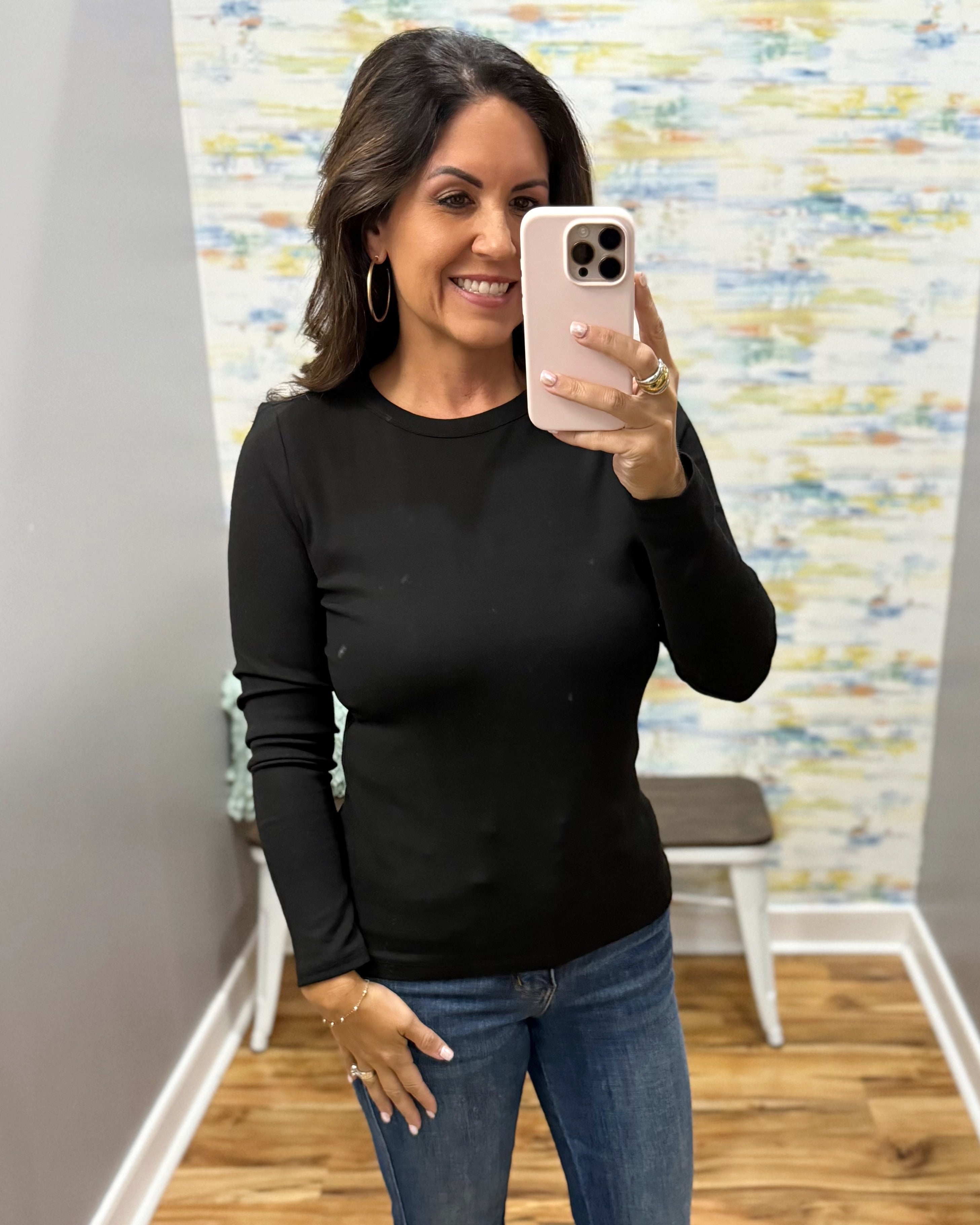 A woman taking a mirror selfie in a fitting room wearing a black long-sleeve crewneck top and blue jeans.
