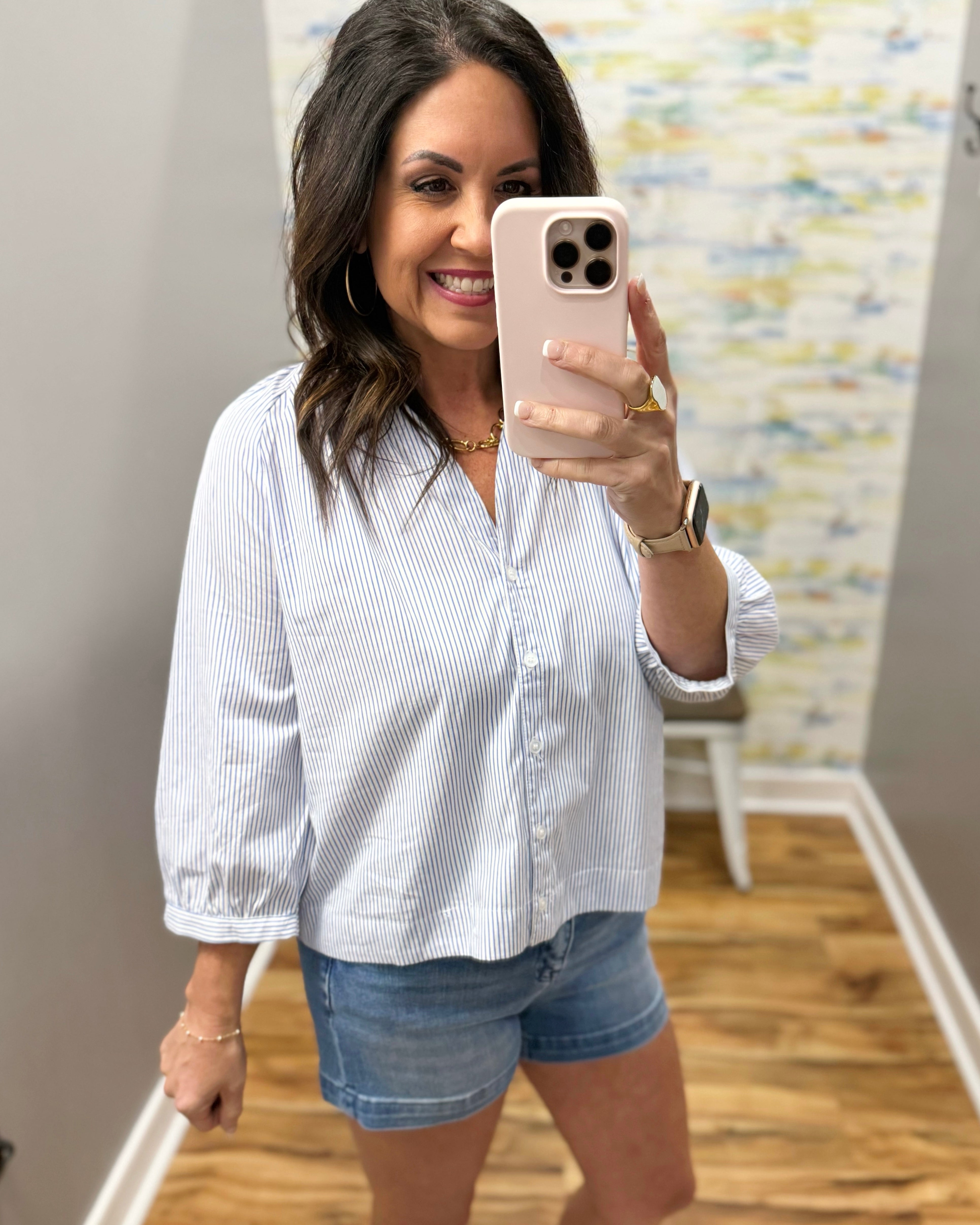 Woman taking a mirror selfie wearing a white striped shirt and denim shorts in a room with a colorful wall.