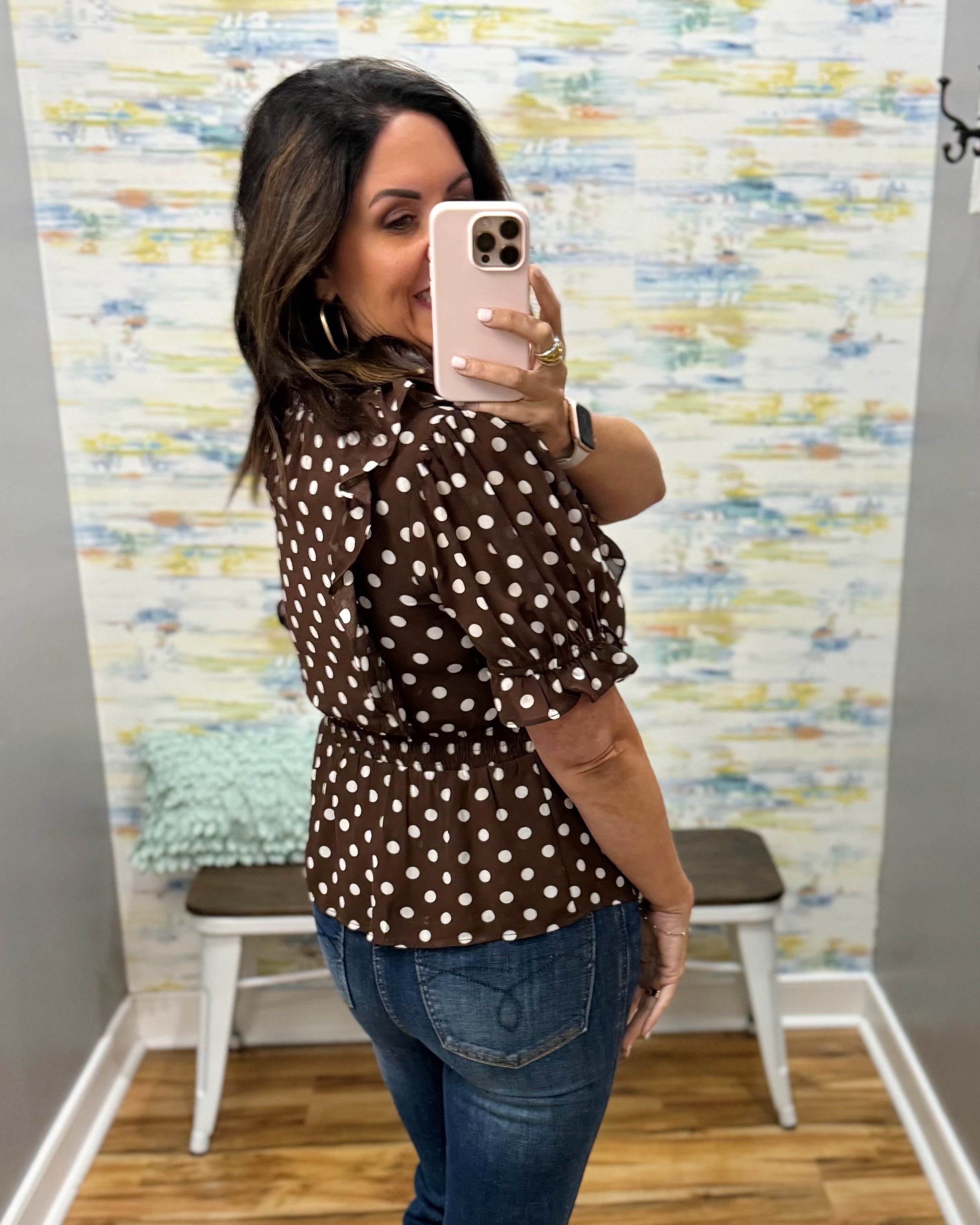 A woman takes a mirror selfie wearing a brown long-sleeve blouse with white polka dots and blue jeans in a fitting room with a patterned wallpaper background.