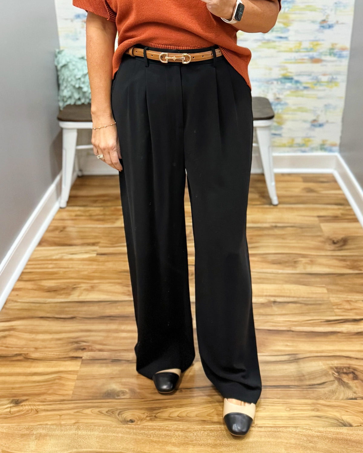 A person wearing black wide-leg pleated trousers with a brown leather belt, paired with an orange top and black-and-white pointed-toe flats, standing on a wood floor.
