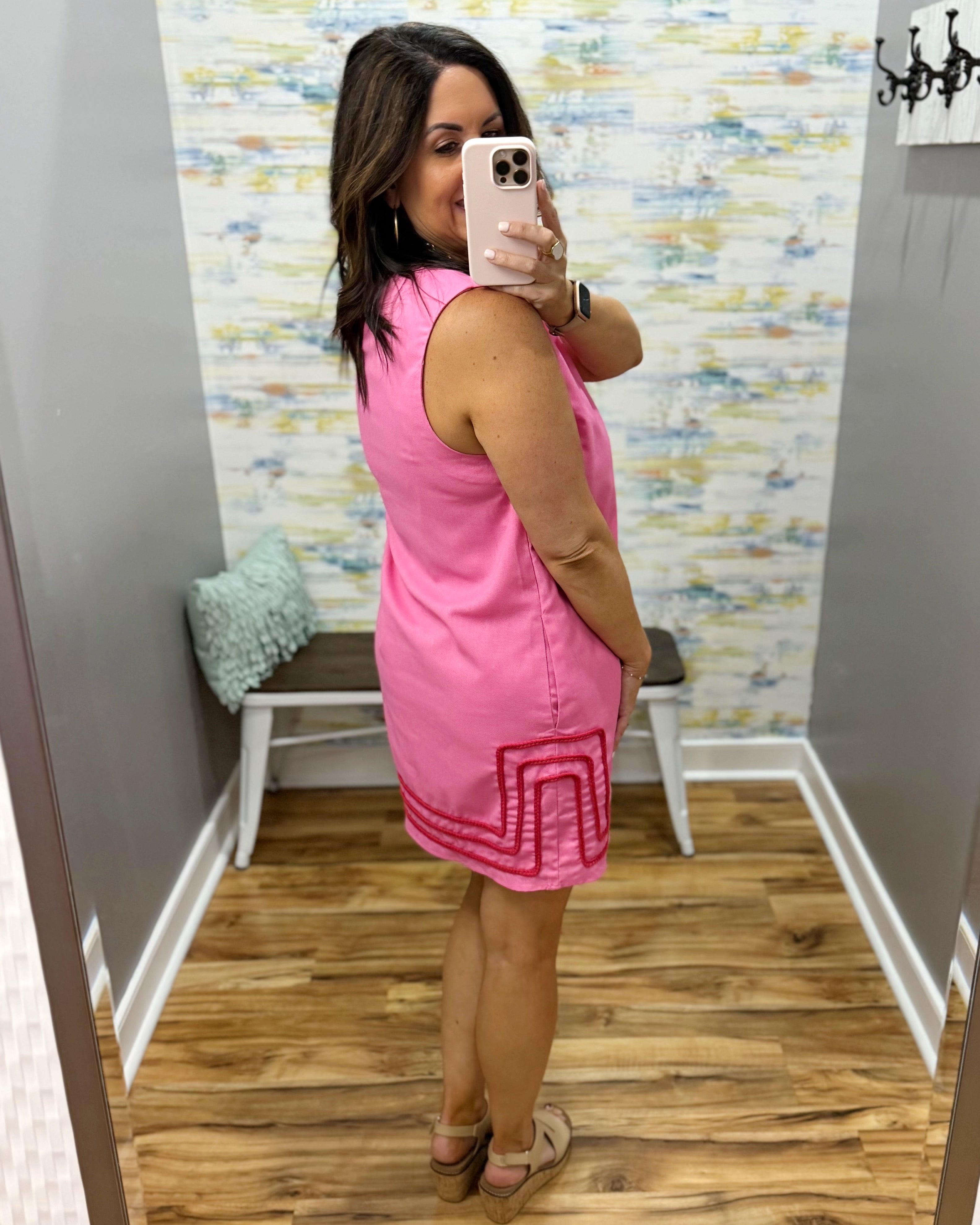 Woman taking a mirror selfie wearing a pink dress with a pattern, standing in a room with a colorful wall and wooden floor.