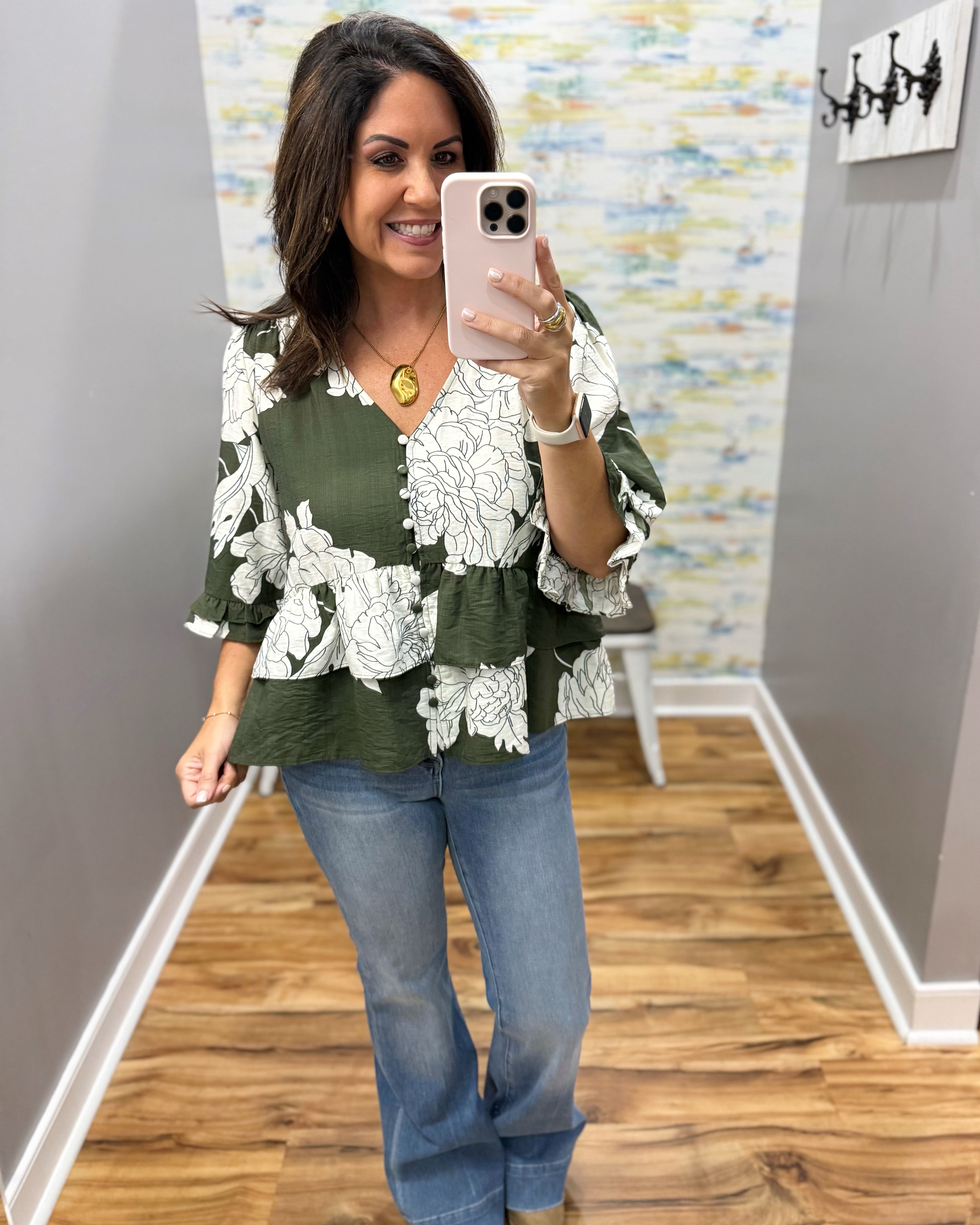 A woman takes a mirror selfie in a boutique showing an olive green and white floral print blouse with ruffled sleeves, paired with blue jeans.