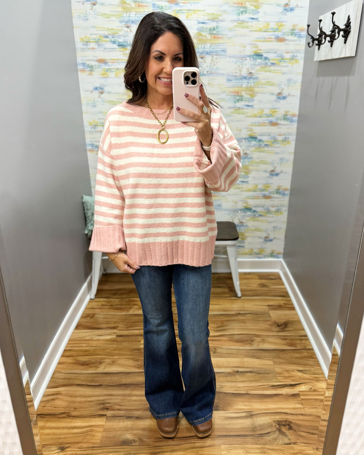 A woman takes a mirror selfie in a fitting room wearing a pink and white striped knit sweater, blue flare jeans, and brown boots.