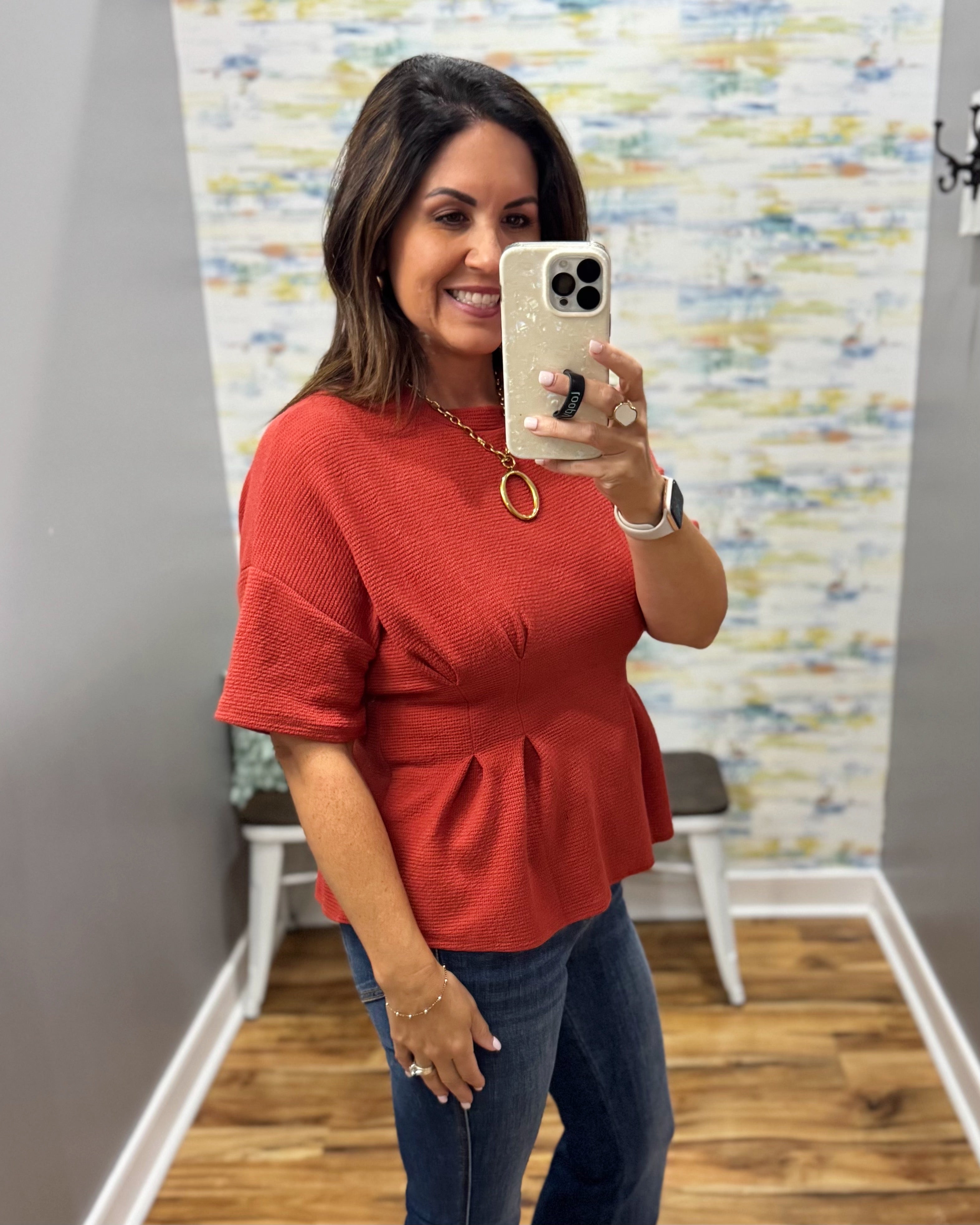 A woman takes a mirror selfie wearing a textured rust-red short-sleeve peplum top and blue jeans, holding a white smartphone.