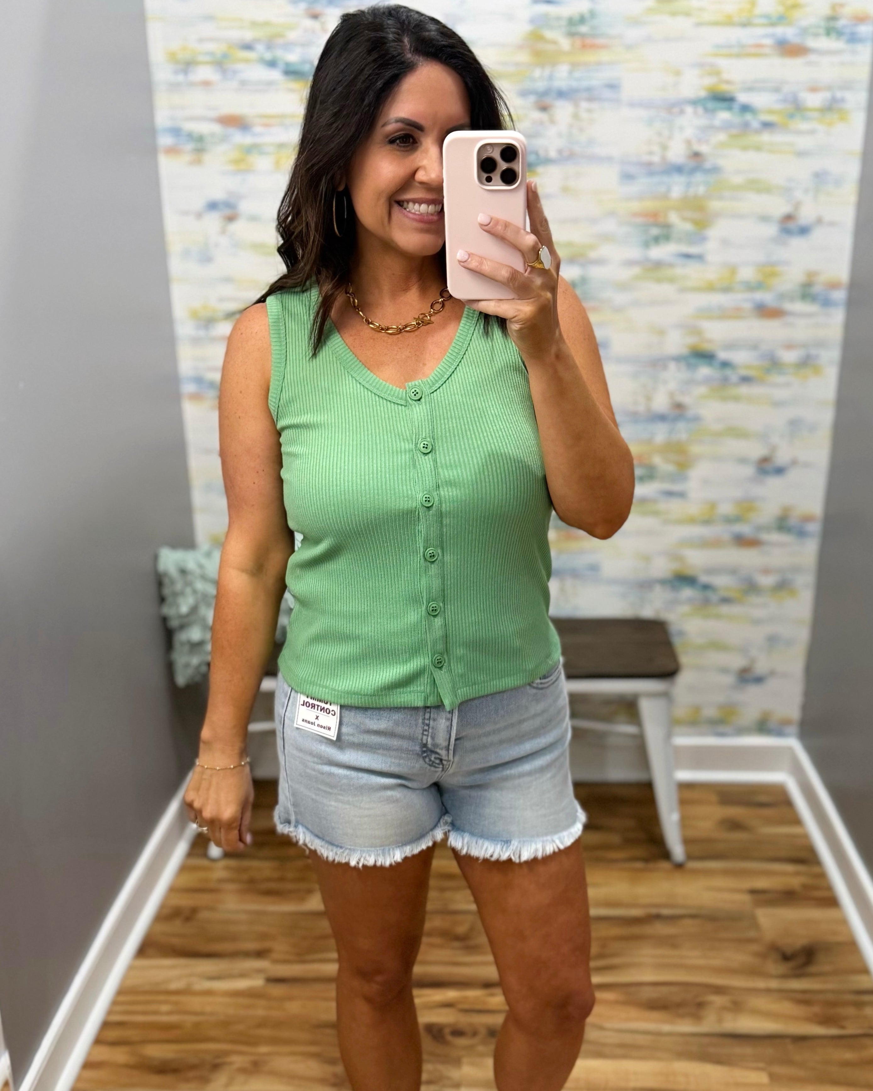 Woman taking a mirror selfie wearing a green sleeveless top and denim shorts in a room with a patterned wall.