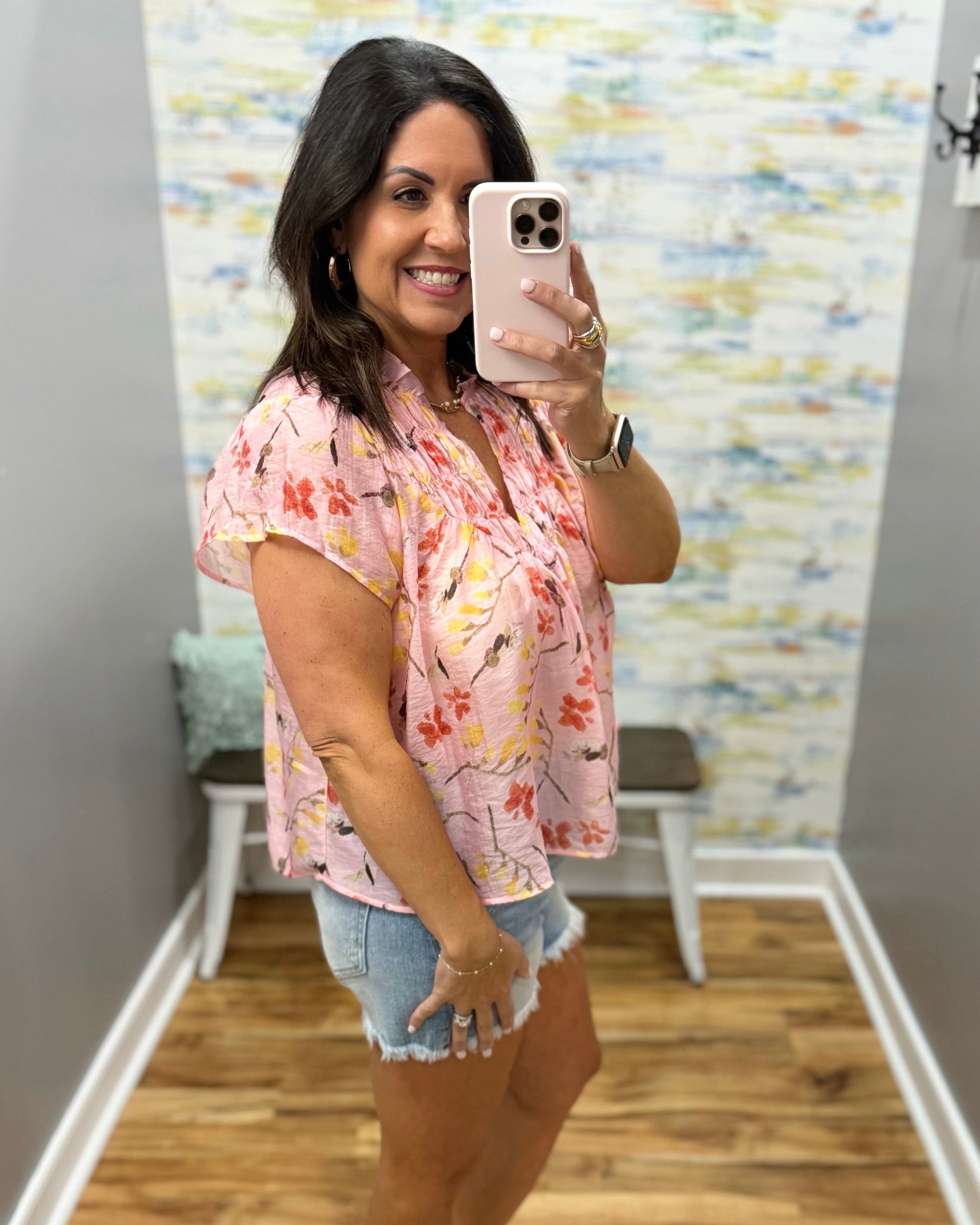 Woman taking a mirror selfie wearing a floral shirt and denim shorts in a room with a colorful wall.