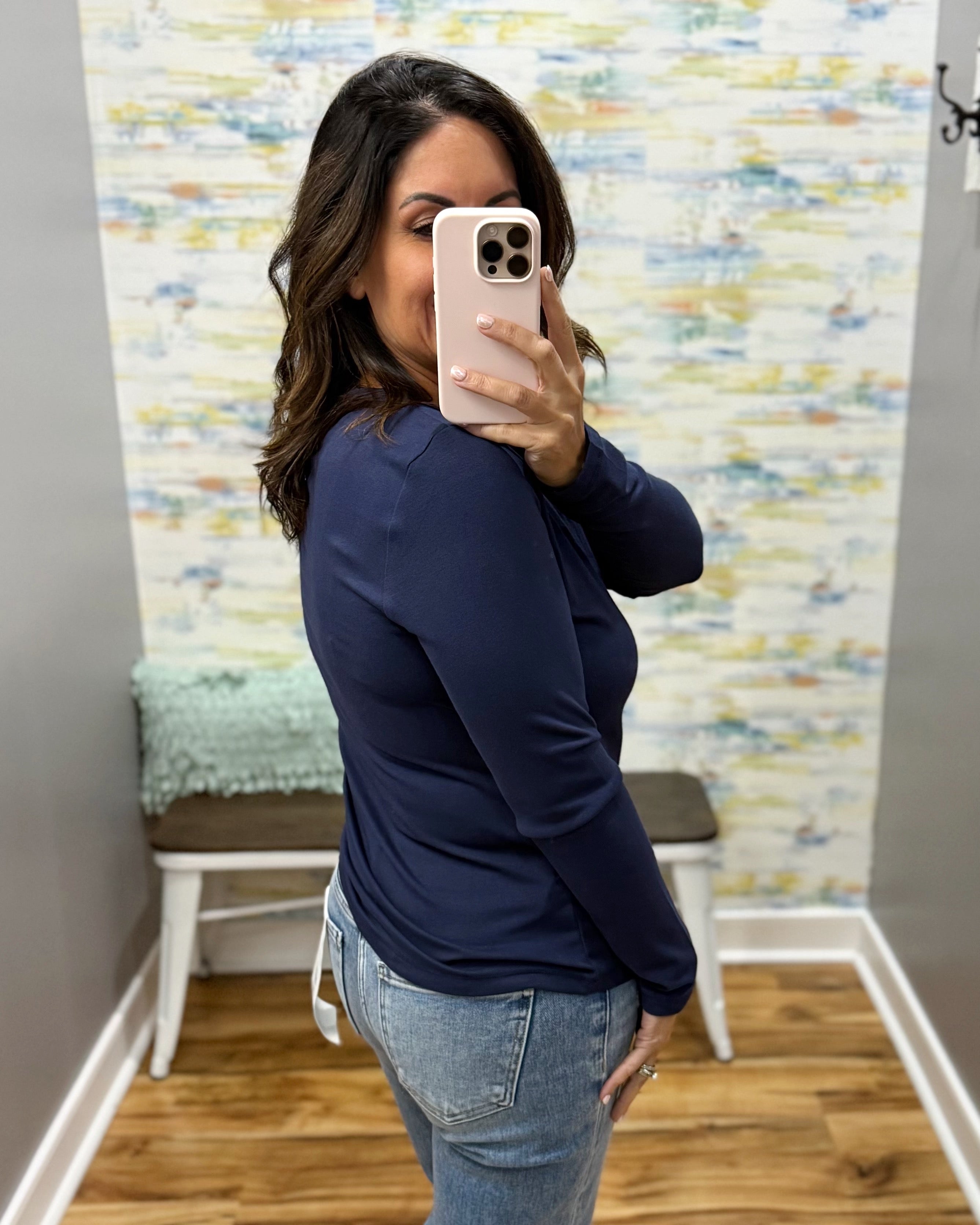 A woman takes a mirror selfie wearing a navy blue long-sleeve top and light-wash blue jeans in a fitting room with a patterned wall and wooden floor.