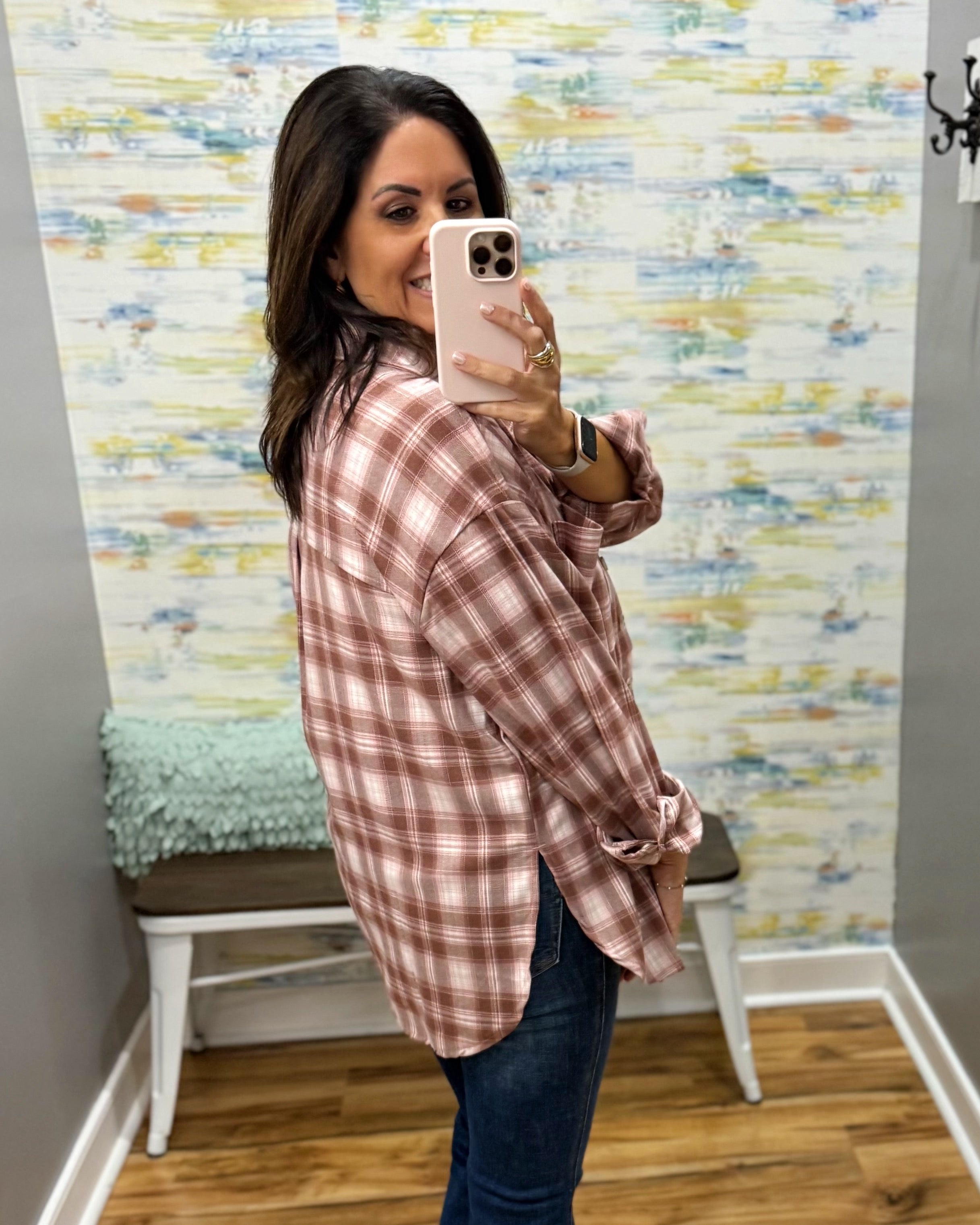 A woman takes a mirror selfie wearing an oversized dusty rose and white plaid button-up shirt with rolled cuffs, paired with dark blue jeans, in a room with patterned wallpaper and a bench.