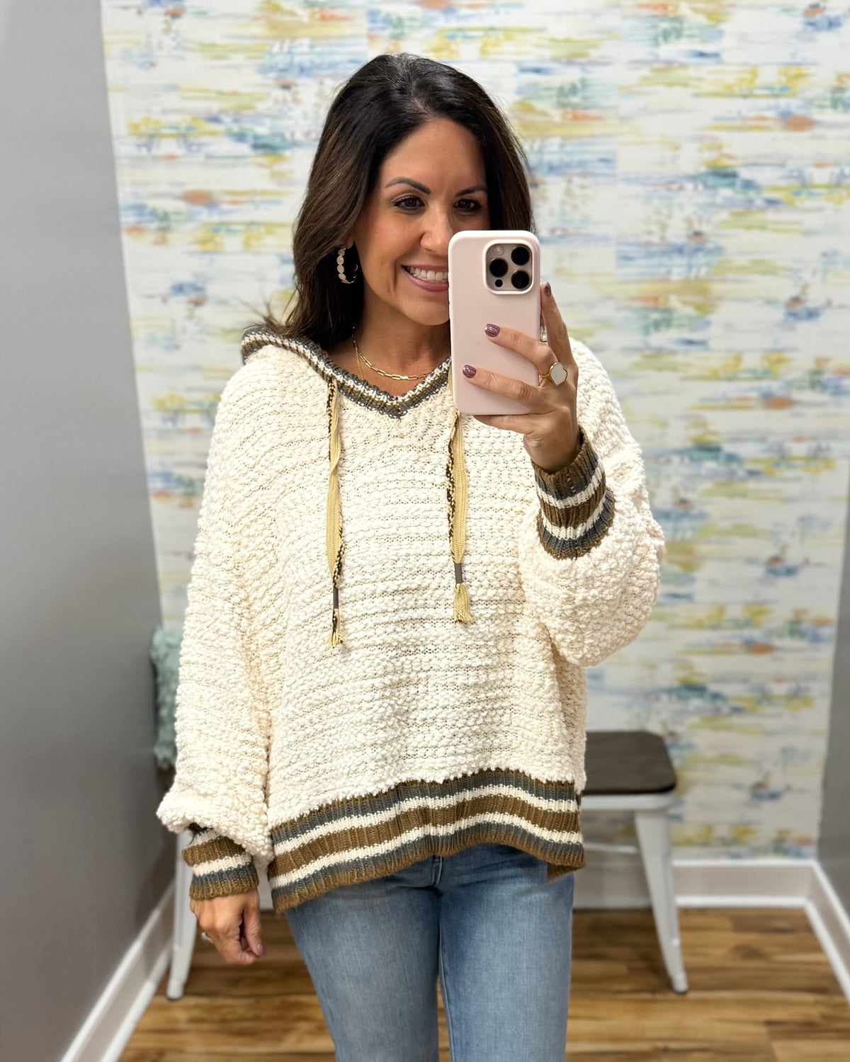 A woman takes a mirror selfie wearing a chunky cream-colored knit hoodie sweater with olive green and brown striped details on the hood, cuffs, and hem, paired with blue jeans.