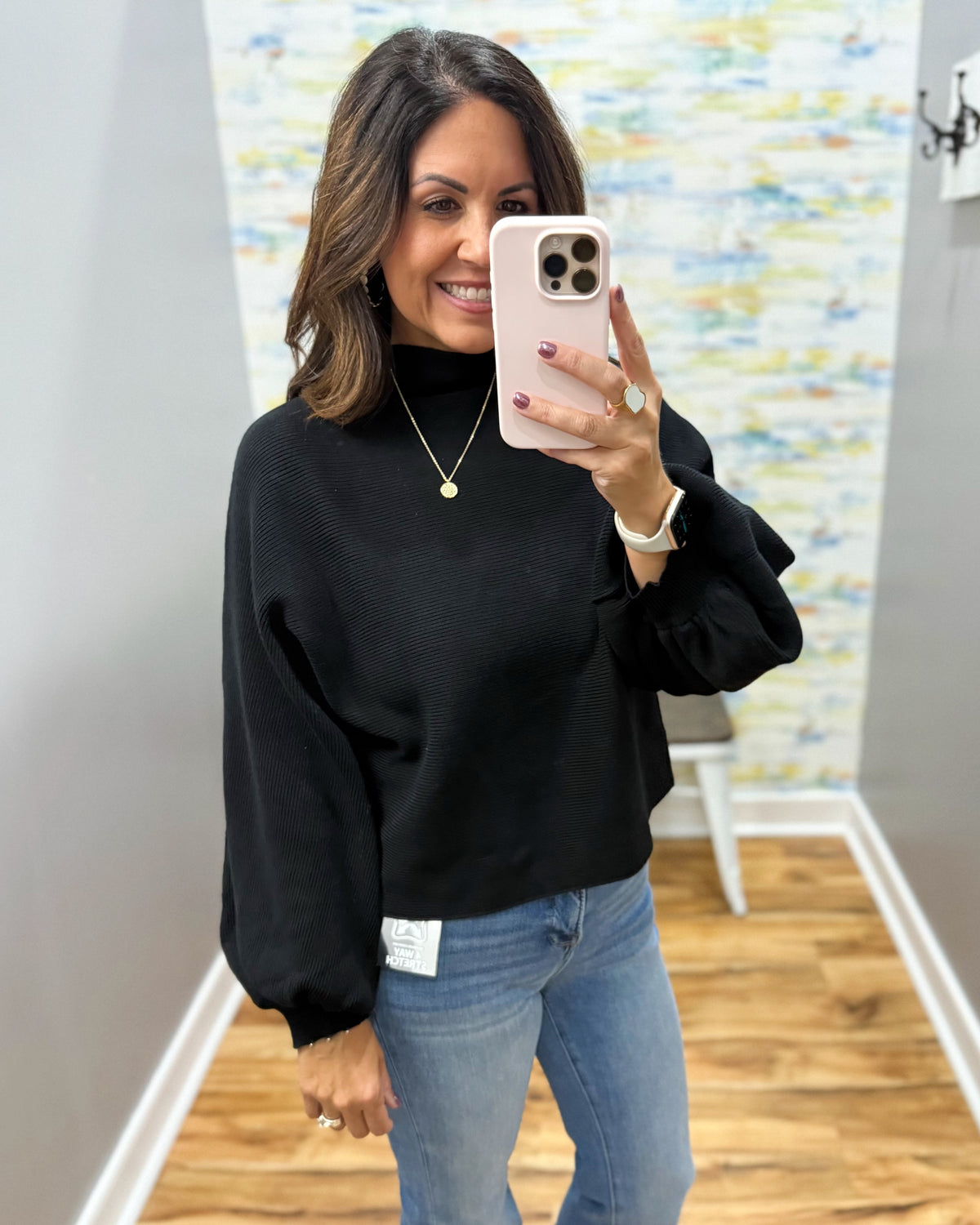 A woman takes a mirror selfie wearing a black long-sleeve mock neck sweater and blue jeans in a home setting with a wood floor and abstract wall art.