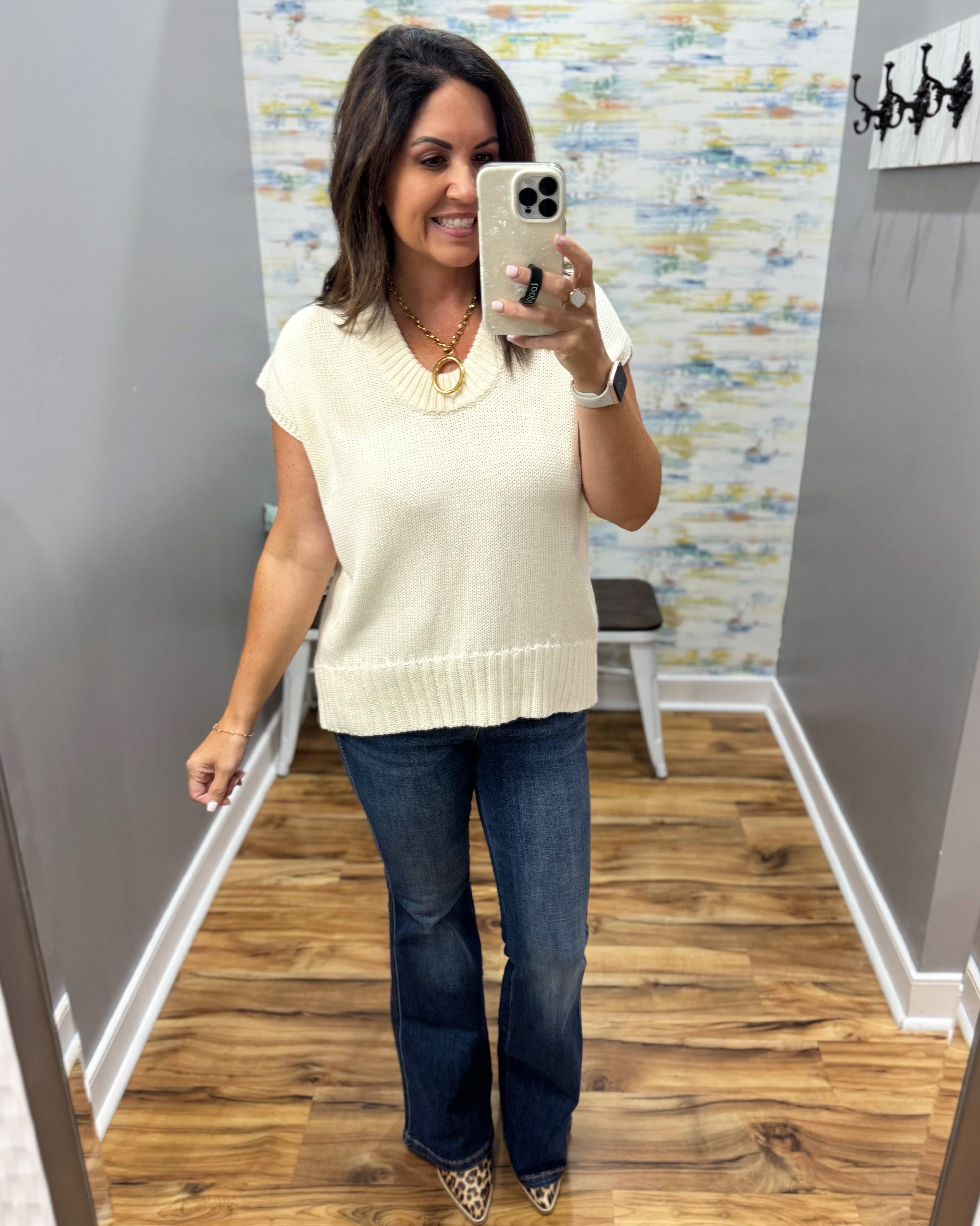 A woman takes a mirror selfie in a fitting room wearing a cream ribbed knit sleeveless sweater vest with a cowl neckline, paired with dark blue flare jeans.