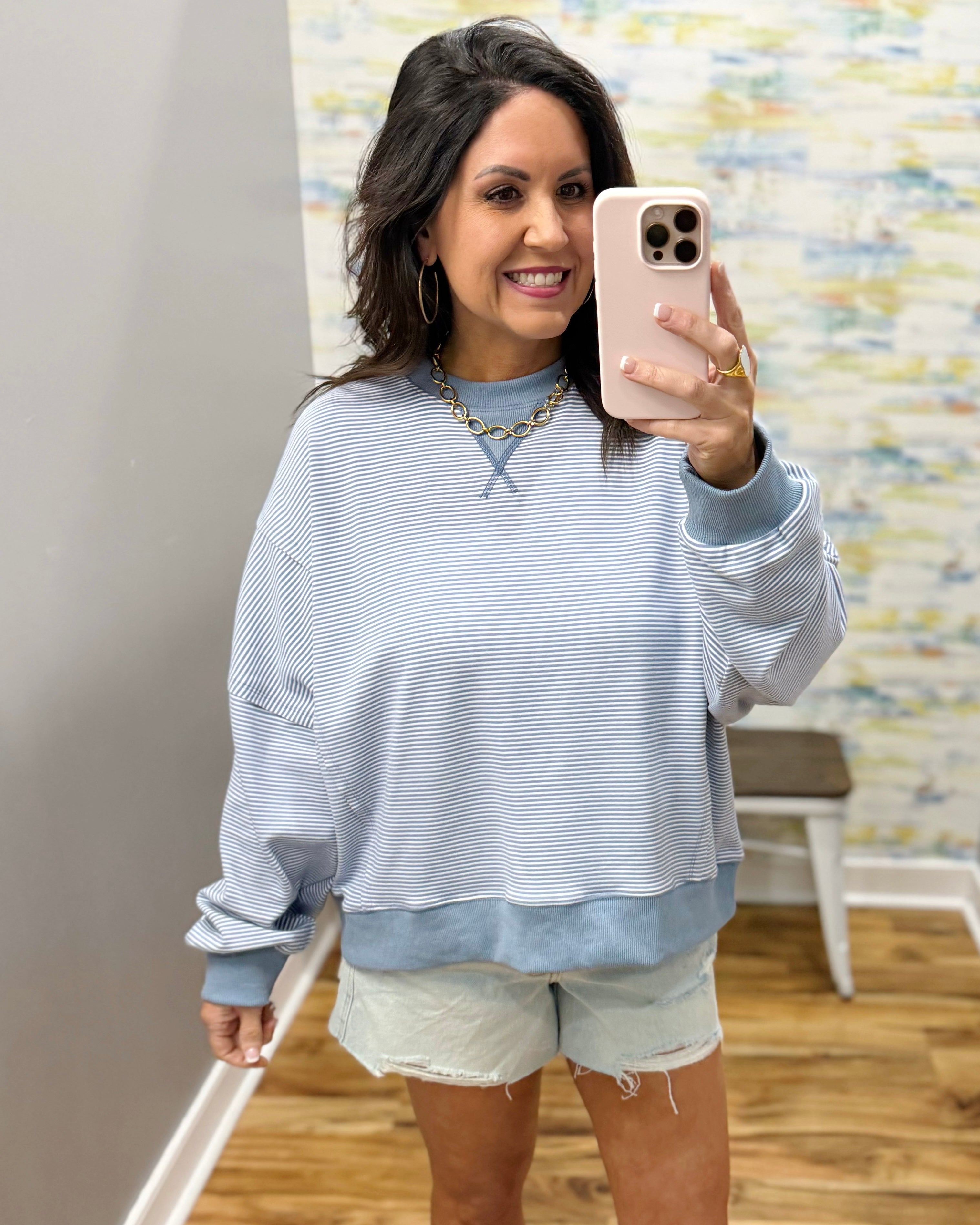 Woman taking a mirror selfie wearing a light blue striped sweater and white shorts in a room with patterned wallpaper.