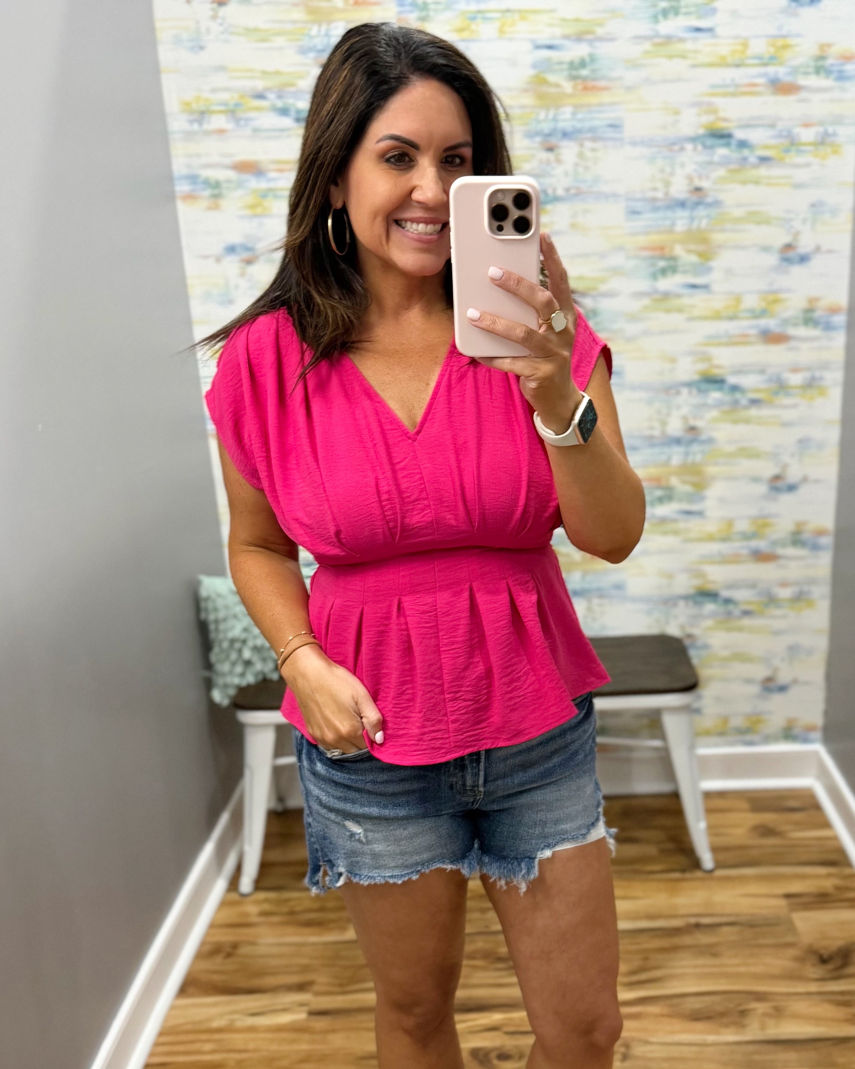 Woman taking a mirror selfie wearing a pink top and denim shorts in a room with a colorful wall.