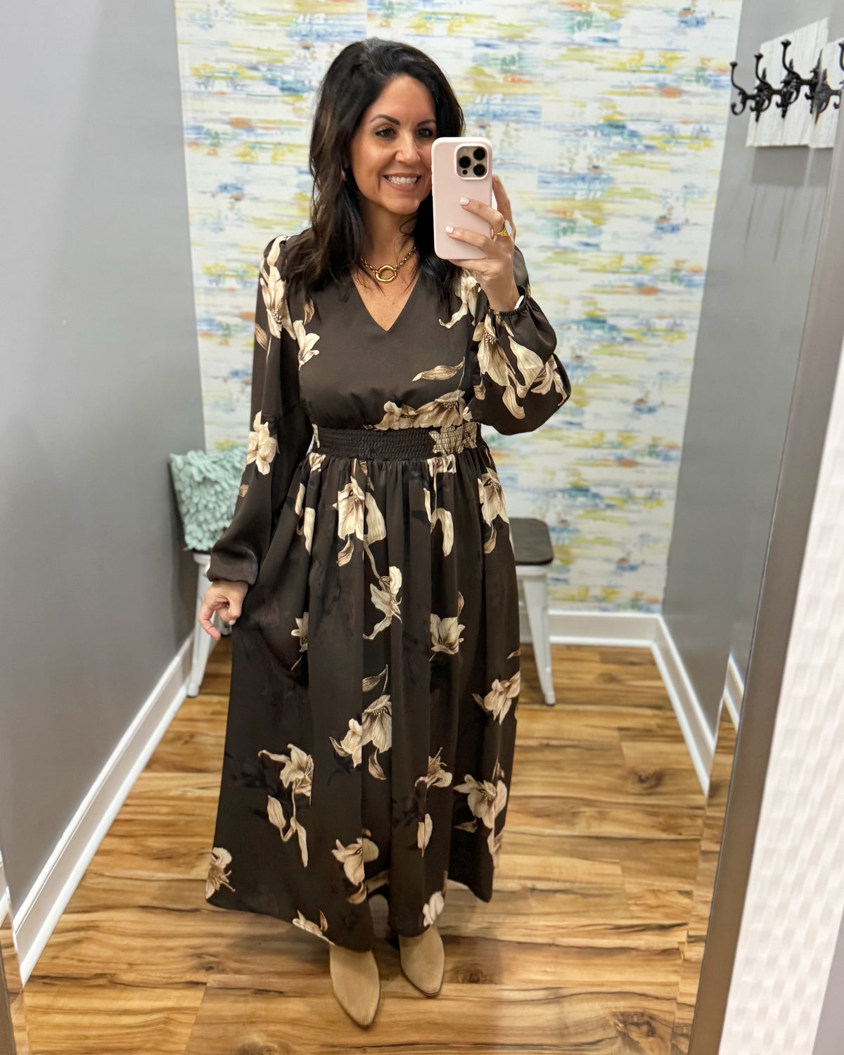 A woman takes a mirror selfie in a boutique fitting room wearing a dark brown long-sleeve maxi dress with a beige floral pattern and a matching waist tie belt.