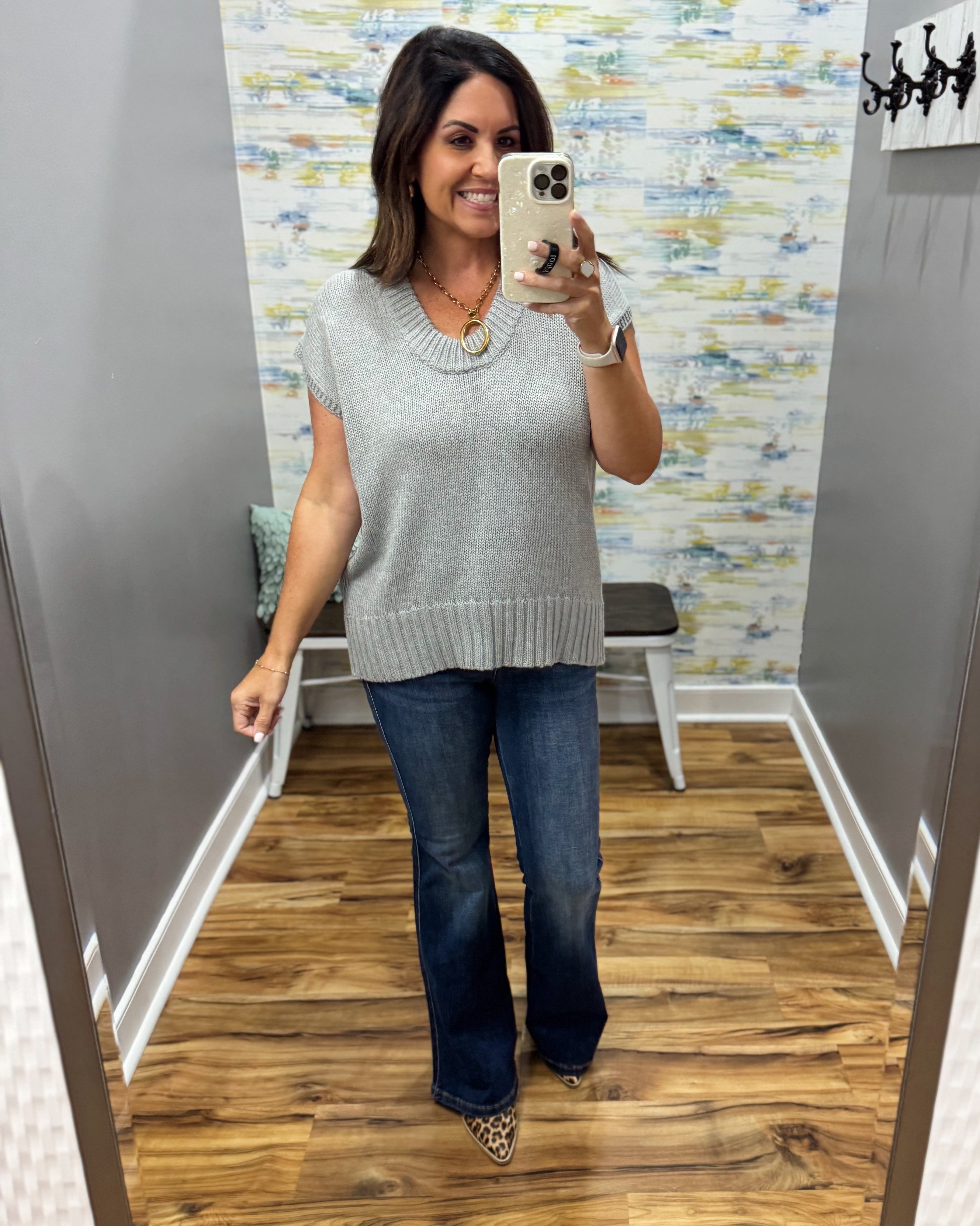 A woman takes a mirror selfie in a fitting room wearing a light gray short-sleeve knit sweater, dark blue flare jeans, and leopard print heels.