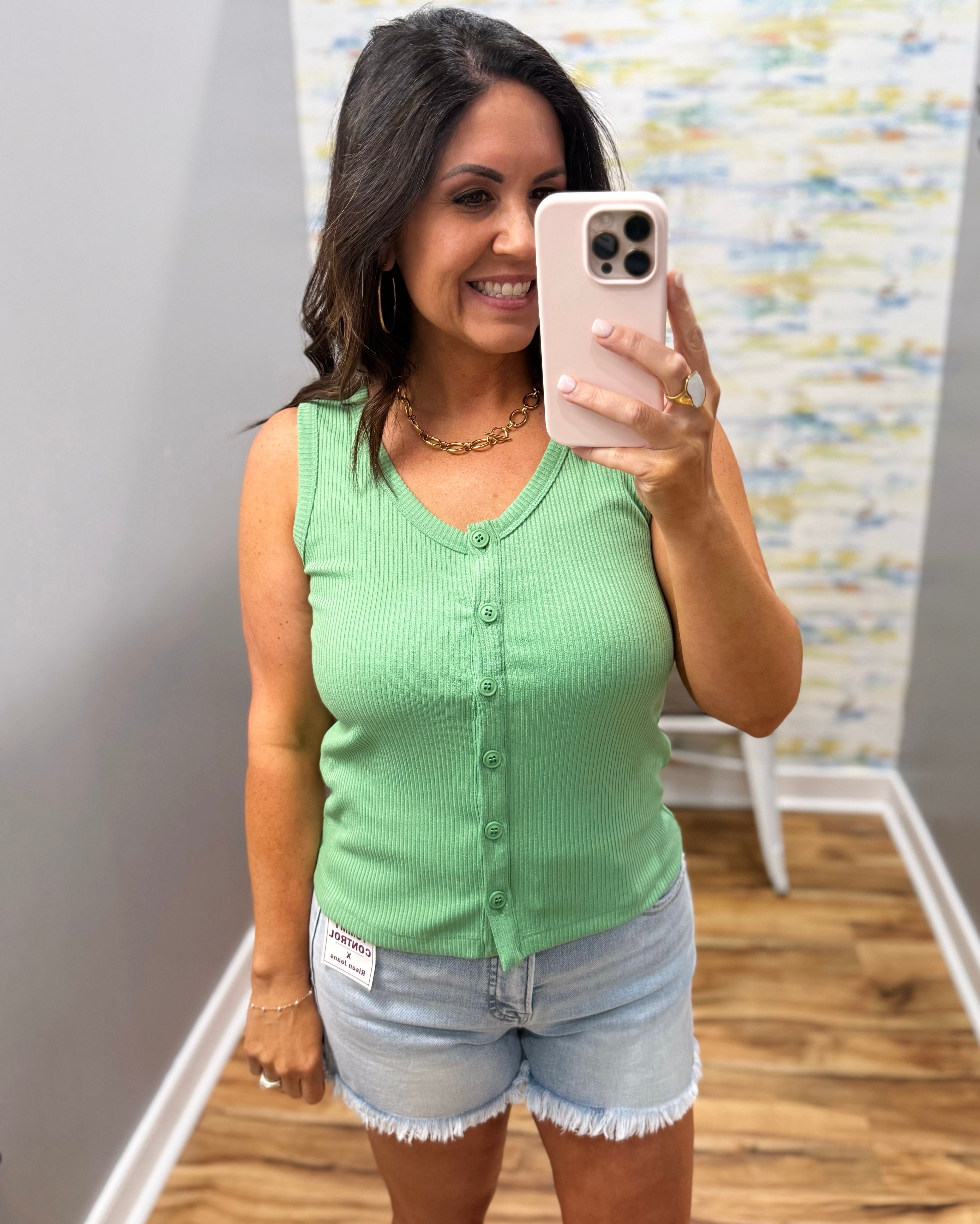 Woman taking a mirror selfie wearing a green sleeveless top and denim shorts.