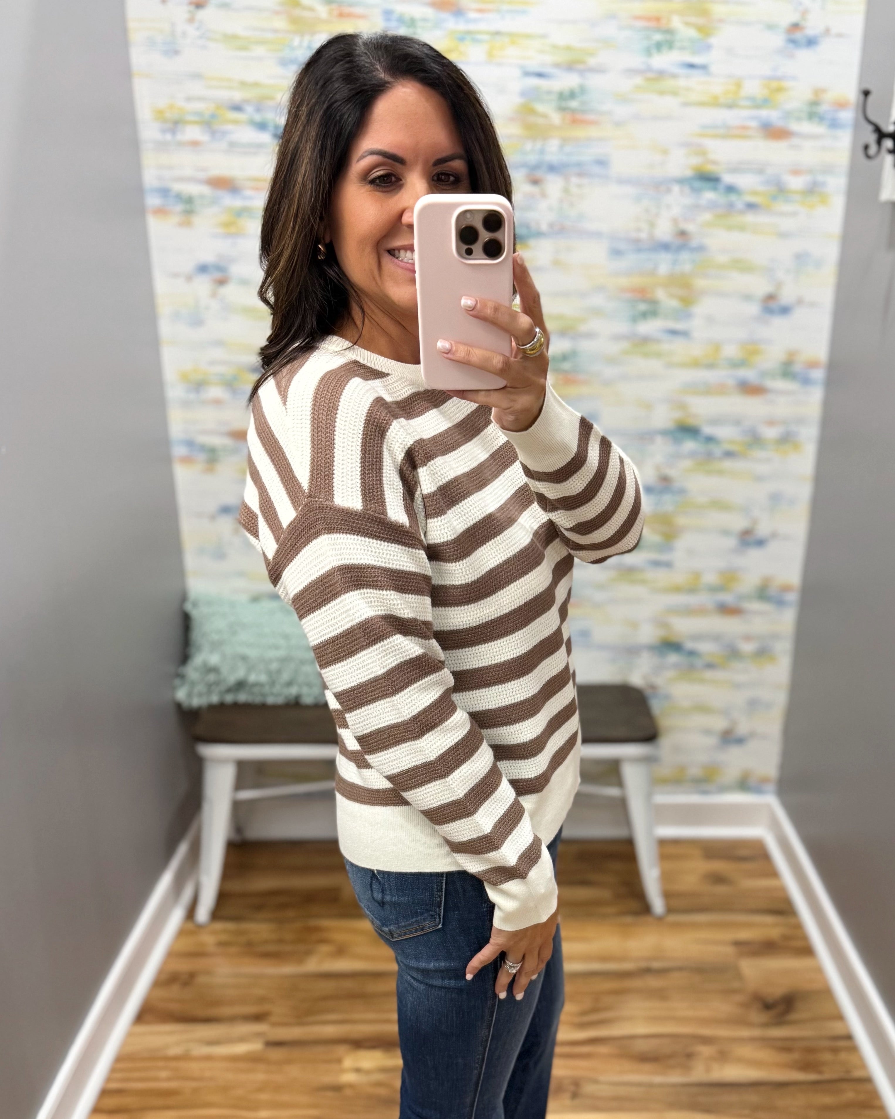A woman takes a mirror selfie wearing a brown and cream striped knit sweater and blue jeans in a home setting with a patterned wall and wood floor.