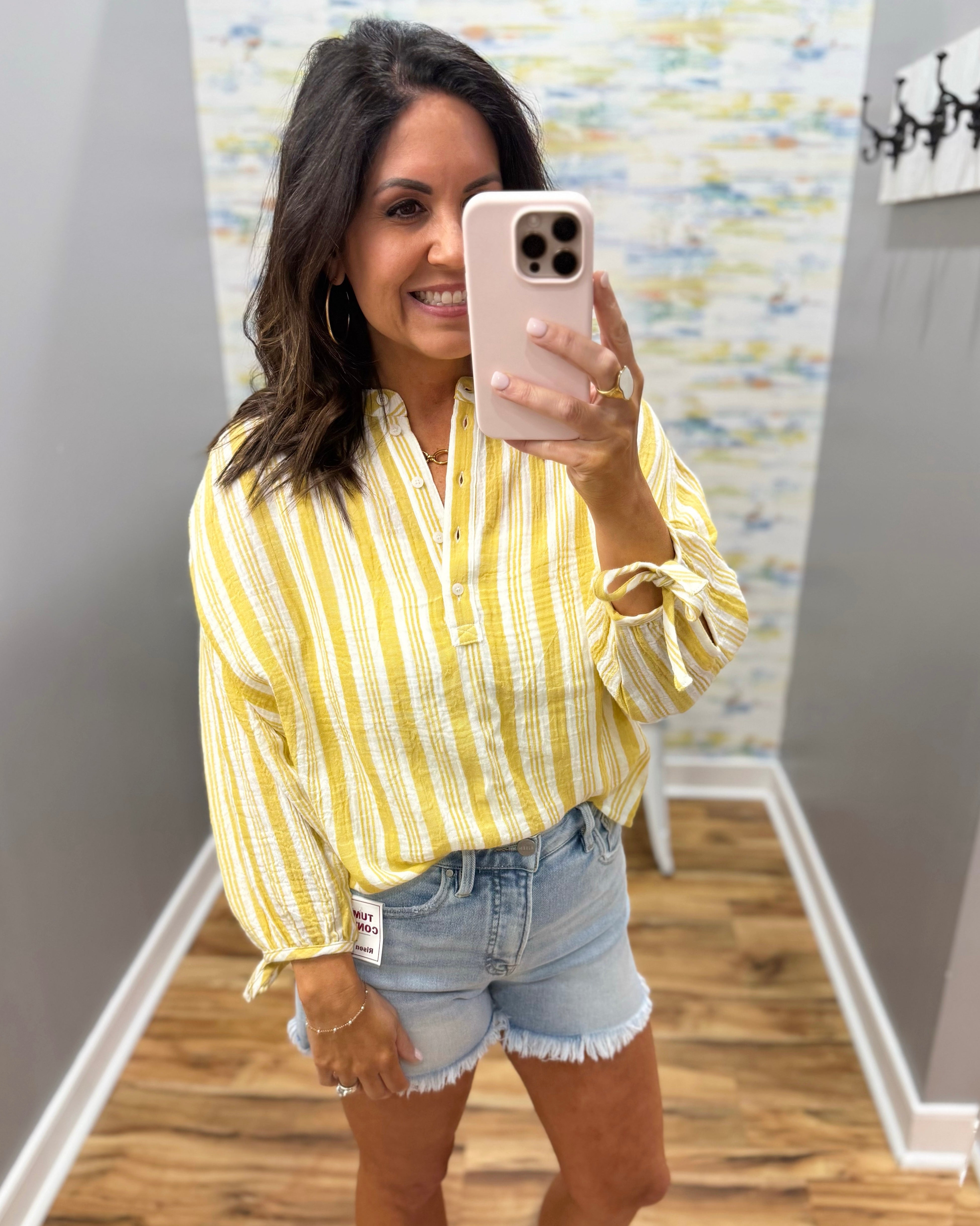 Woman taking a mirror selfie wearing a yellow striped shirt and denim shorts.