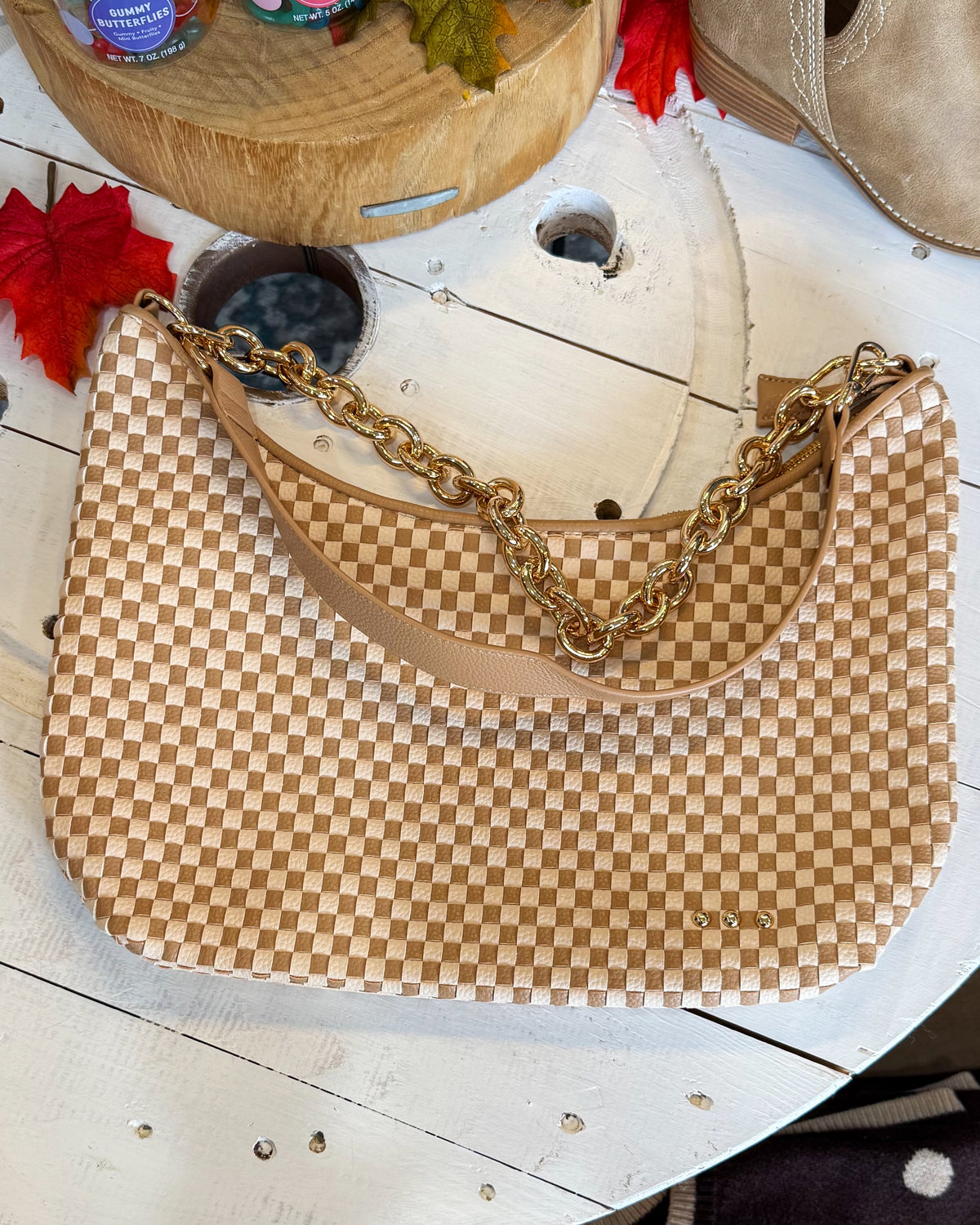 A beige and tan checkerboard shoulder bag with a gold chain strap, displayed on a rustic white wooden table.