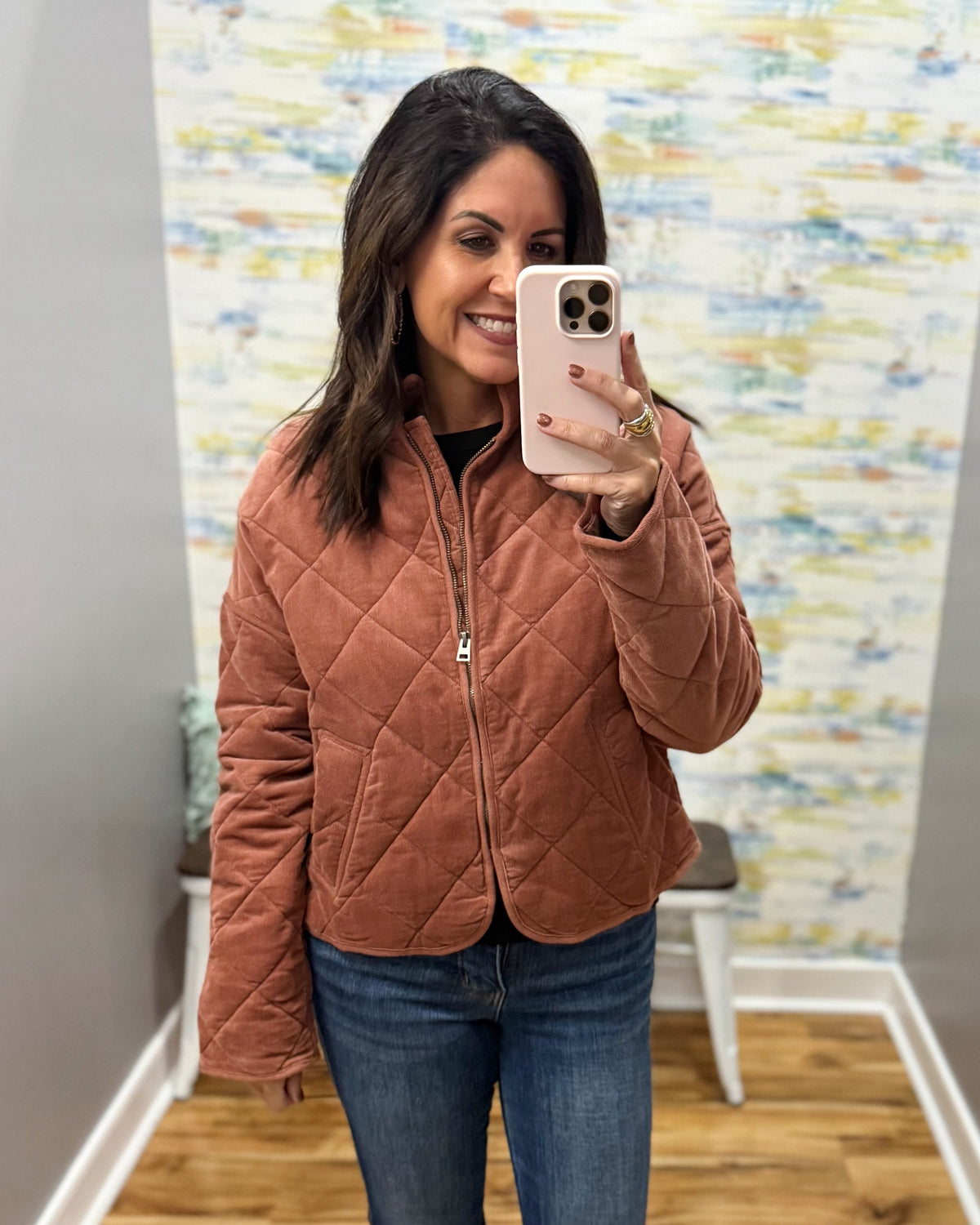 A woman takes a mirror selfie wearing a rust-colored quilted zip-up jacket and blue jeans in a fitting room with a patterned wall and wood floor.