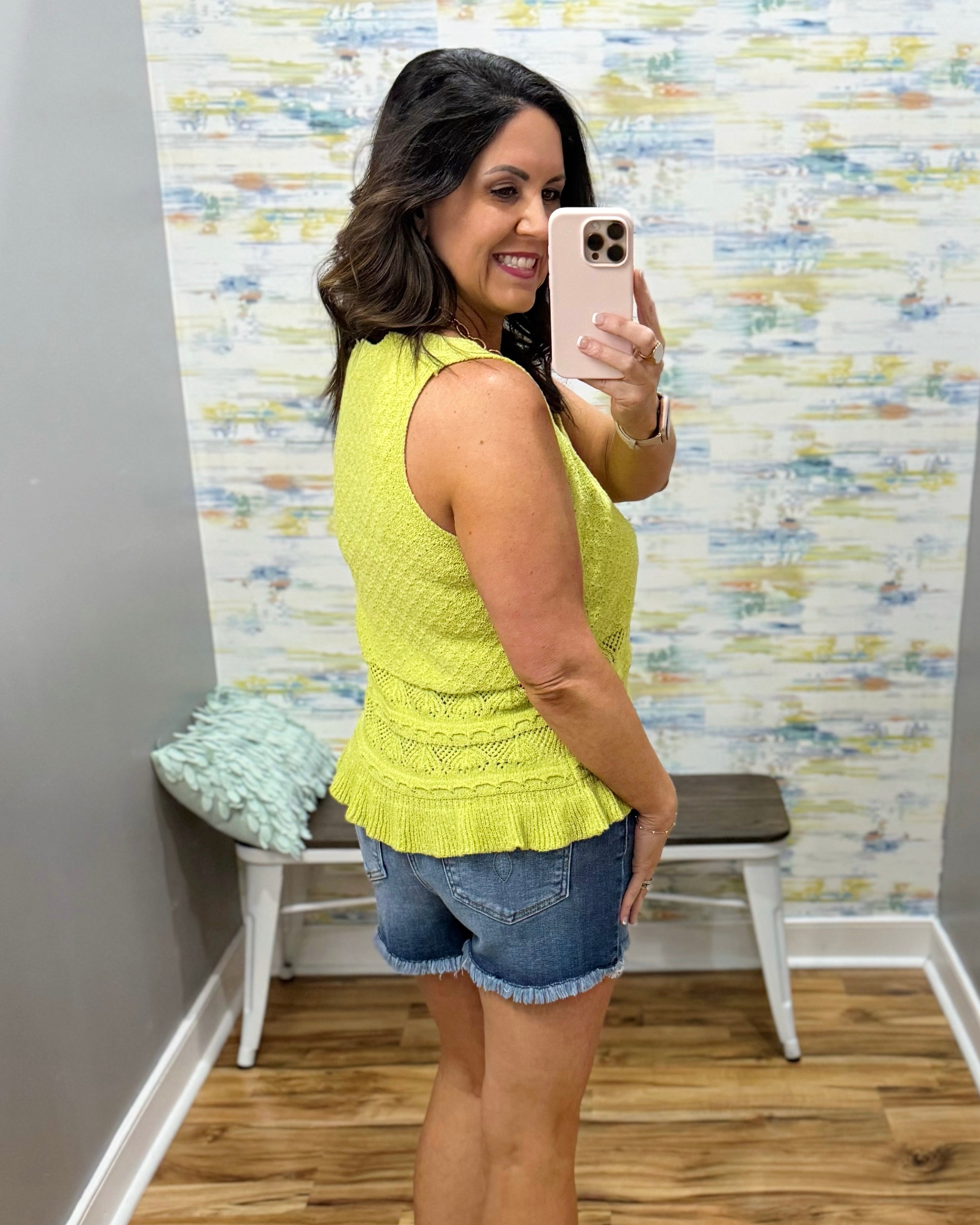 Woman taking a mirror selfie wearing a yellow top and denim shorts in a room with patterned wallpaper.