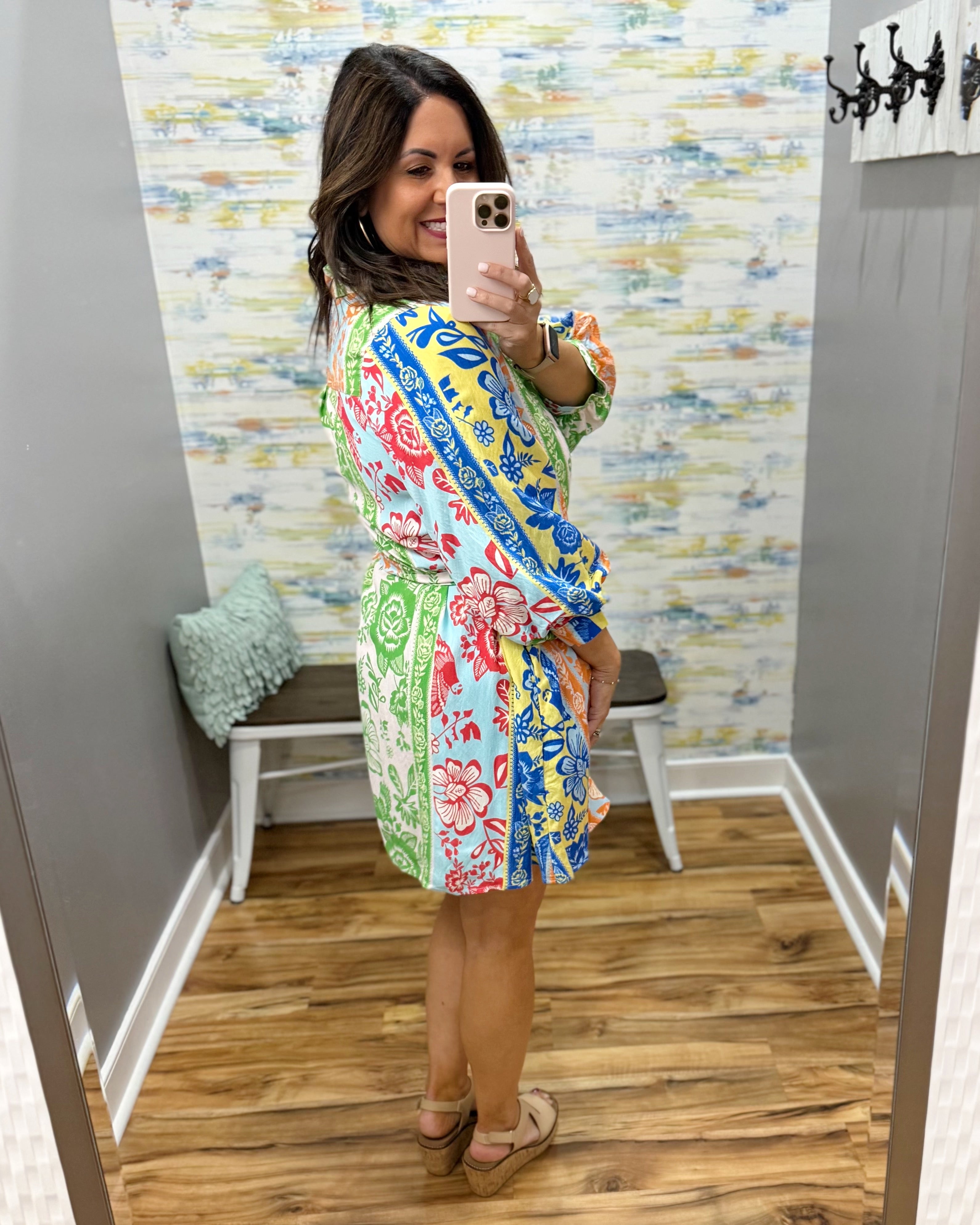 Woman taking a mirror selfie wearing a colorful floral dress in a room with patterned wallpaper.