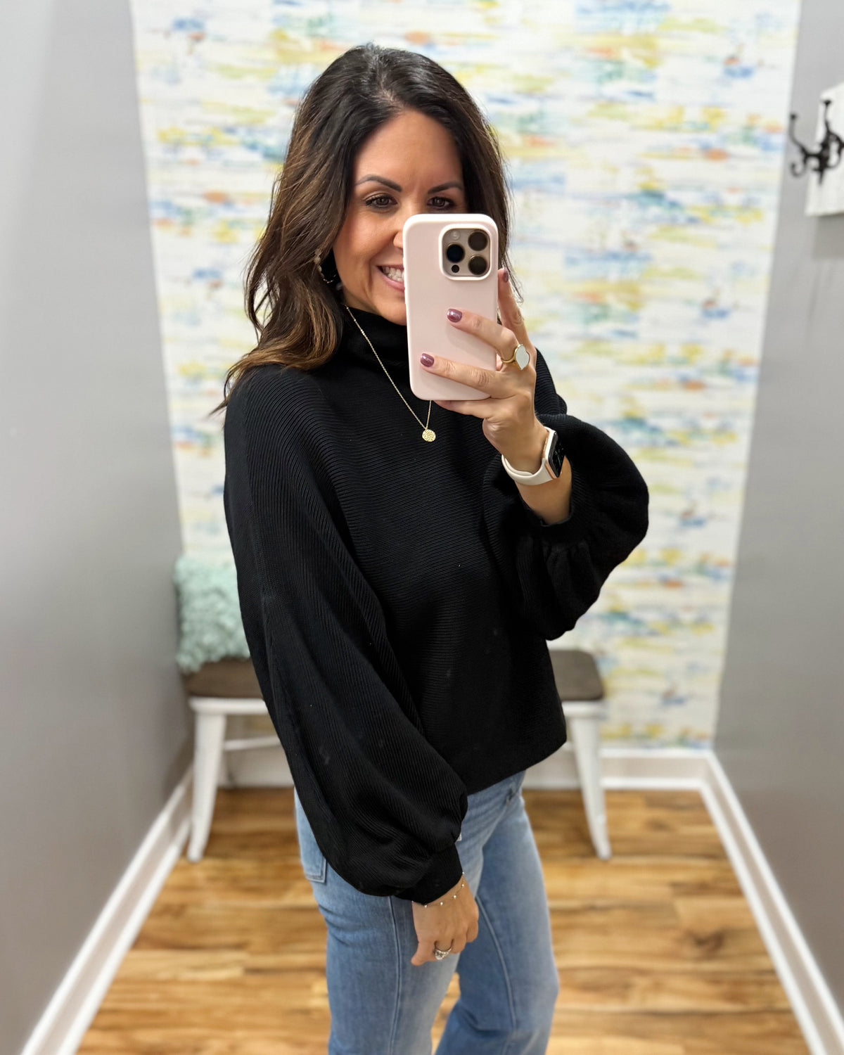 A woman takes a mirror selfie wearing a black long-sleeve knit top and light blue jeans in a fitting room with wood floors and a patterned wall.
