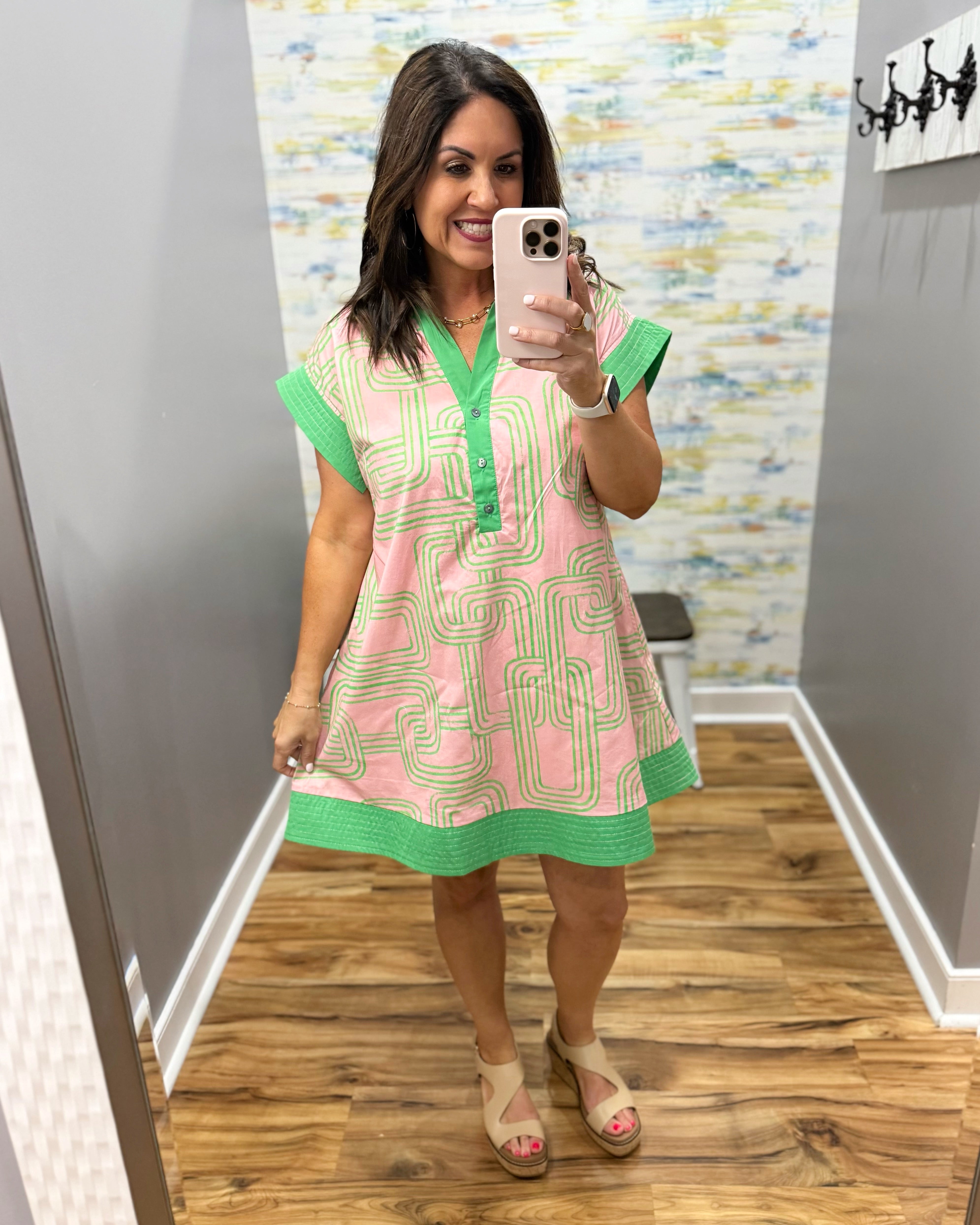 Woman taking a mirror selfie wearing a pink and green dress in a room with patterned wallpaper.
