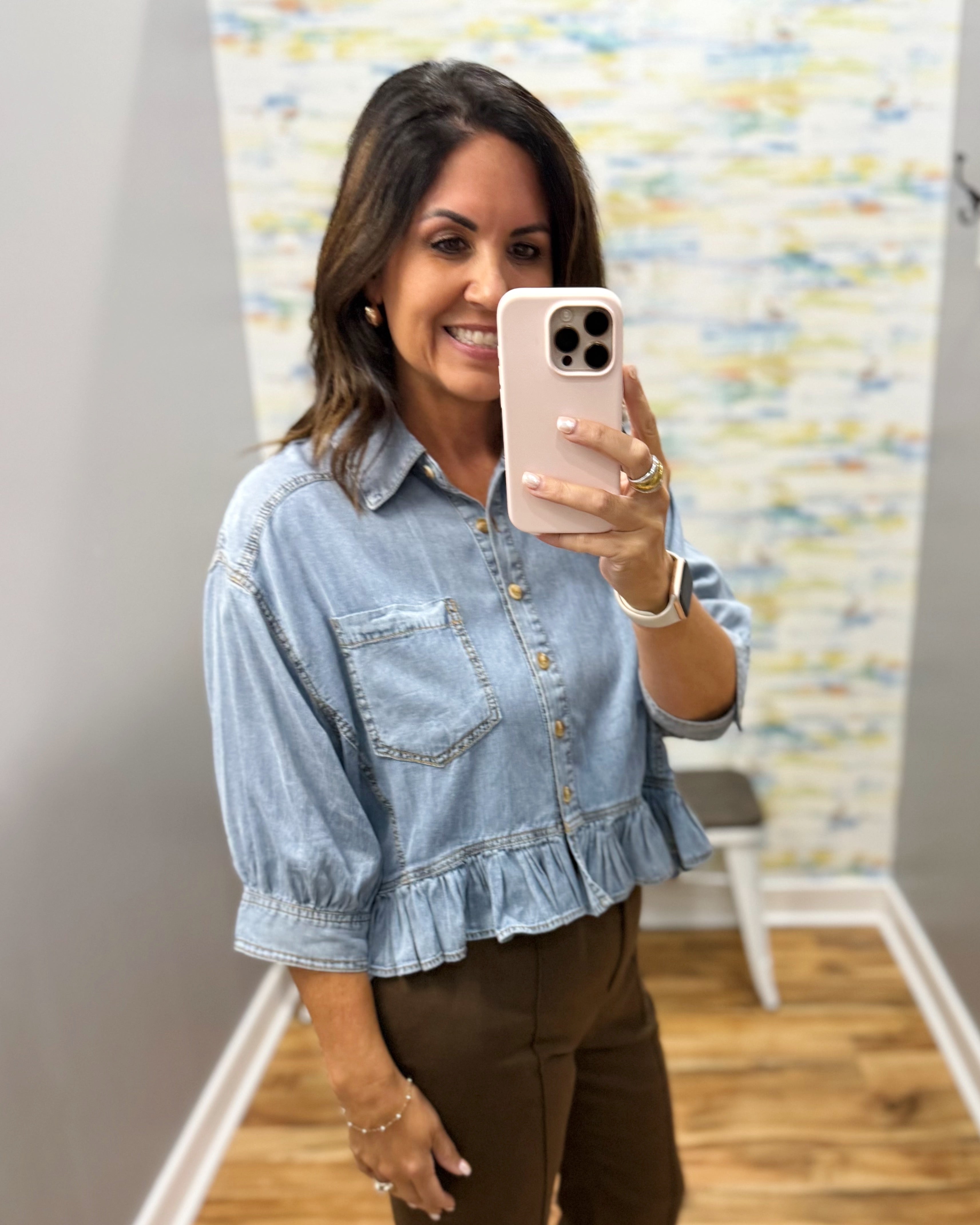 A woman takes a mirror selfie wearing a light blue denim button-up shirt with a ruffled hem and brown pants in an indoor setting.