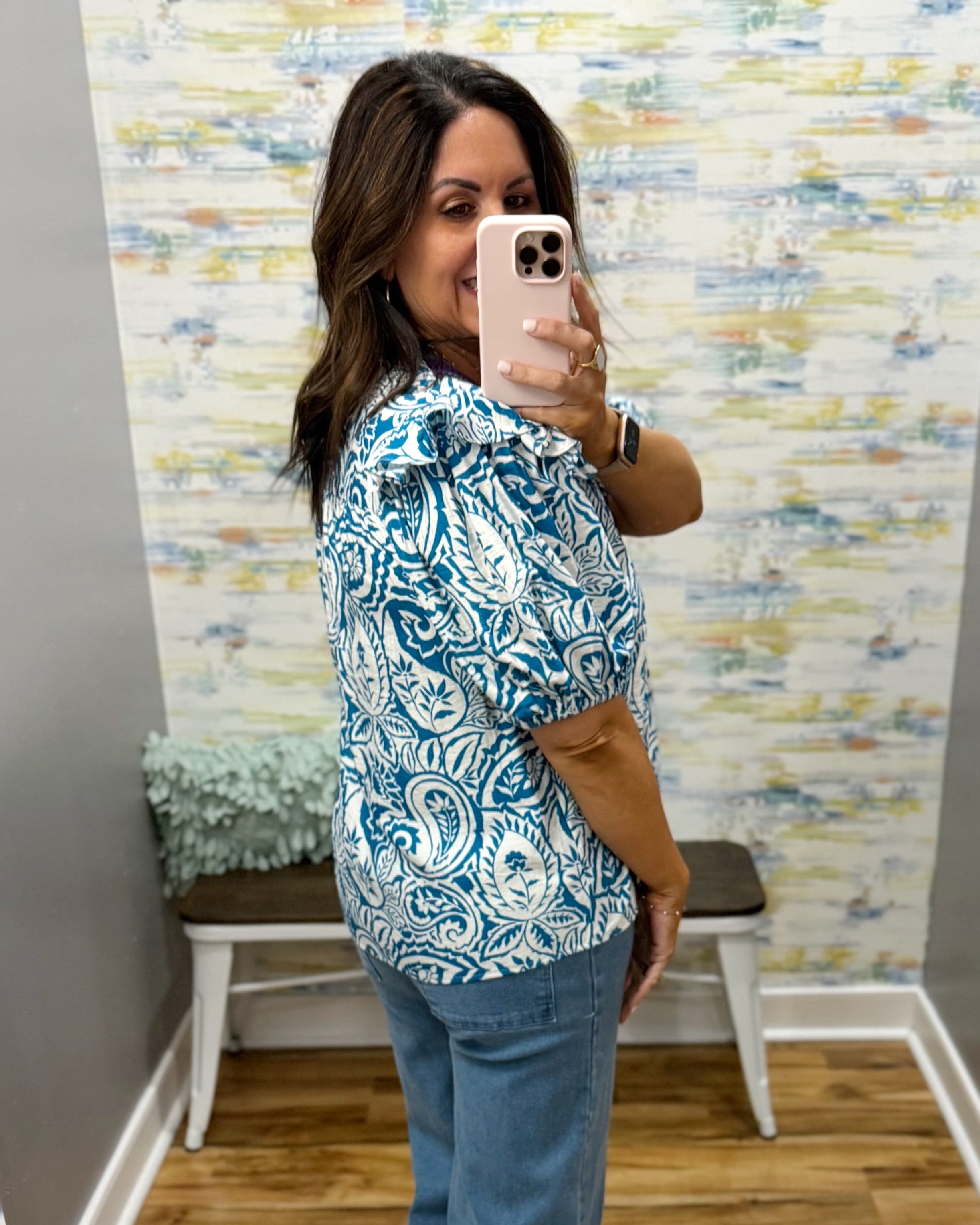 Woman taking a mirror selfie wearing a blue and white patterned blouse and jeans in a room with colorful wall art.